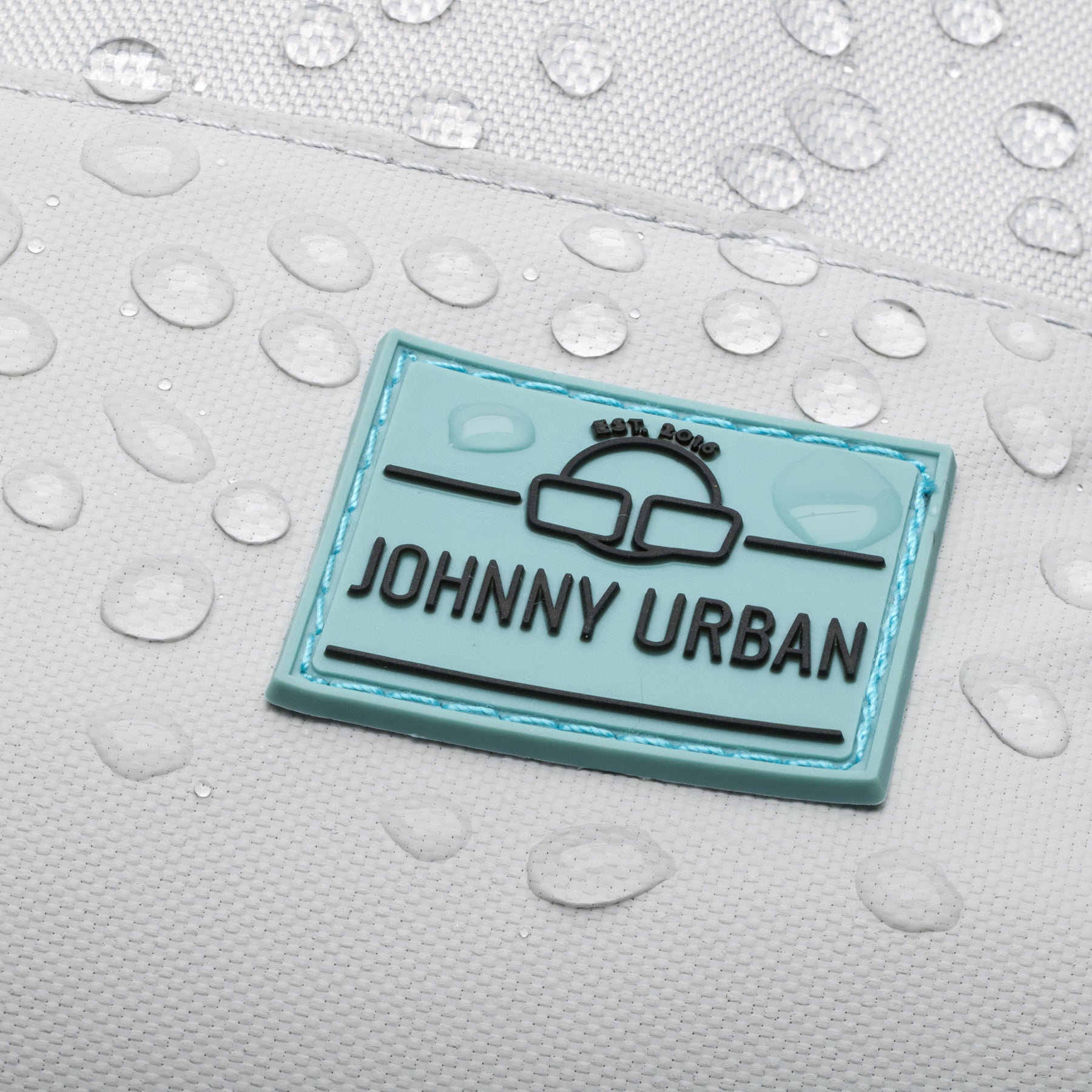 Blaues Johnny Urban Logo auf grauem wasserdichtem Rucksack mit Wassertropfen