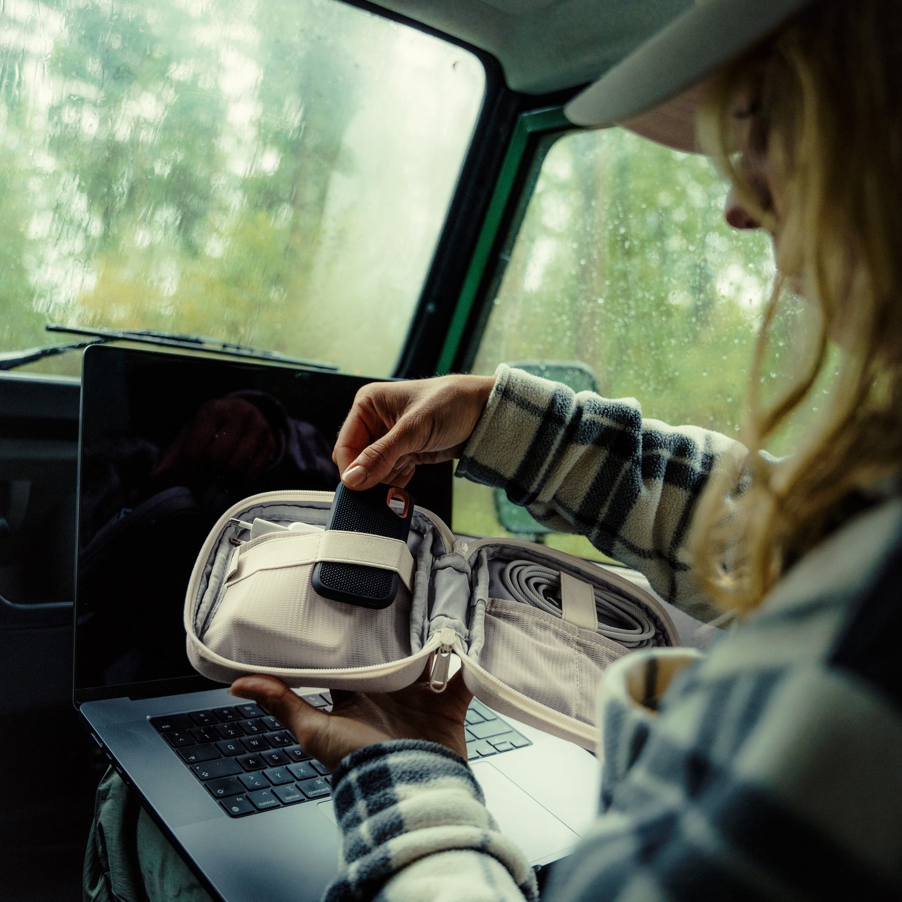 Person in kariertem Hemd nutzt den Tech Organizer 'Ted' zum Verstauen eines Geräts neben einem Laptop im Auto