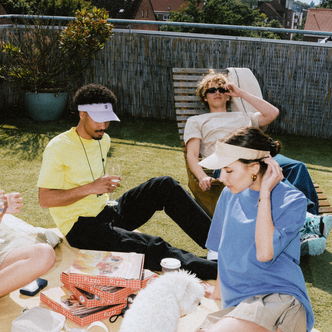 Drei Personen entspannen im Garten bei Sonnenlicht, zwei tragen Sandfarbene Sun Visors 'Terry', eine trinkt aus einem Glas