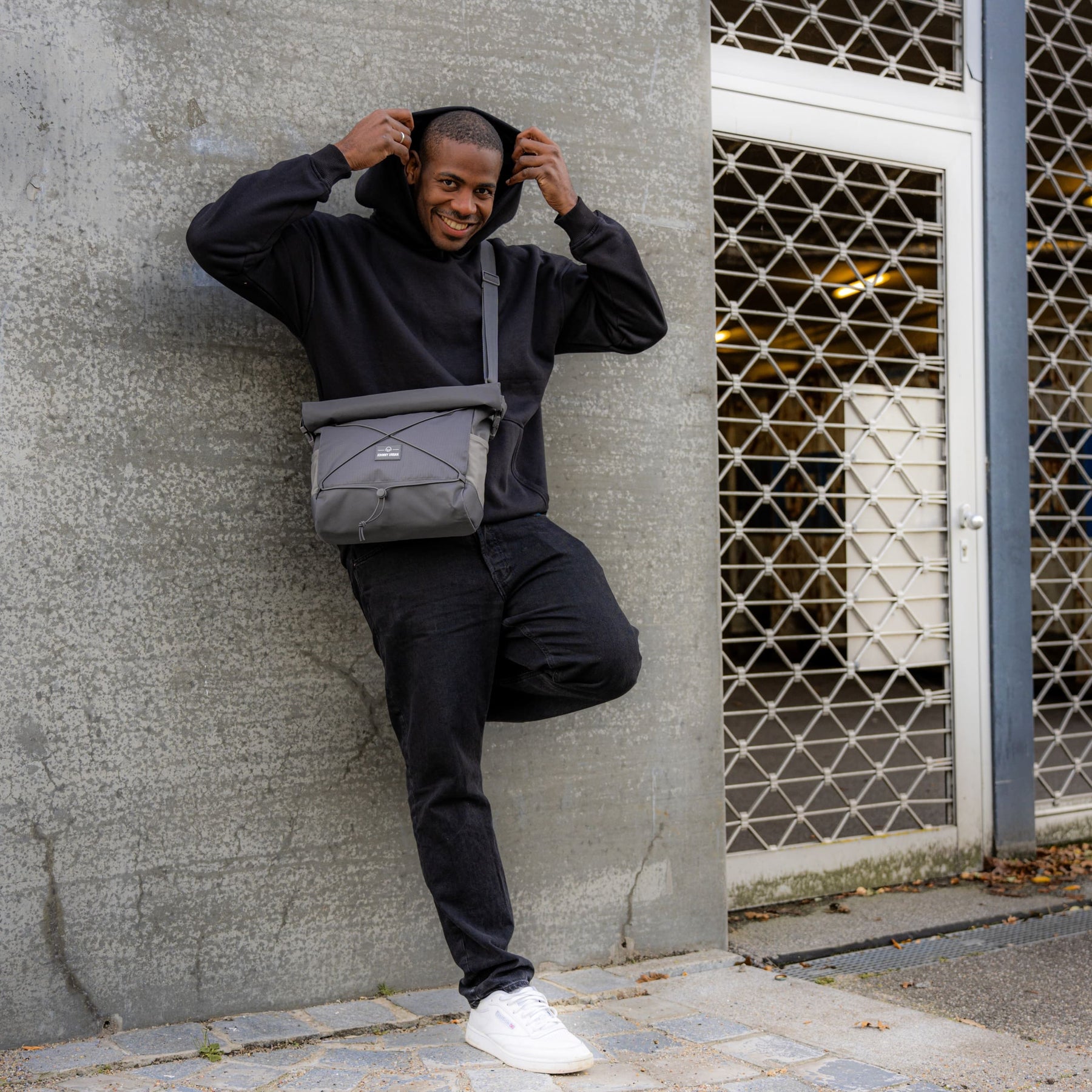 Mann lehnt an Betonwand, trägt schwarze Kapuzenjacke, schwarze Hose, weiße Schuhe und graue Umhängetasche 'Dylan' von Johnny Urban