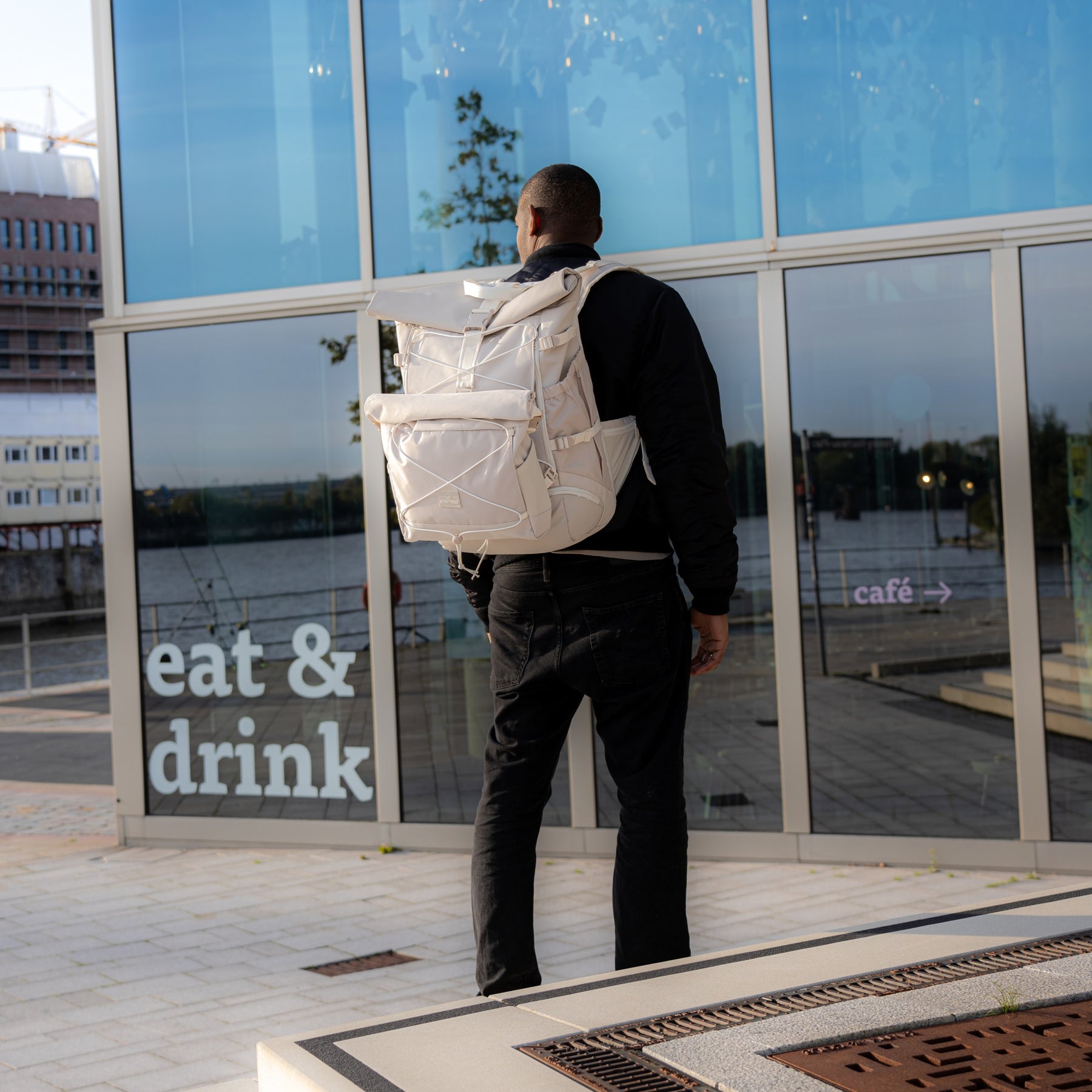 Rückansicht eines Mannes mit schwarzer Jacke und umhängetasche 'Dylan' in beige vor Glasfassade mit Schriftzug 'eat & drink'