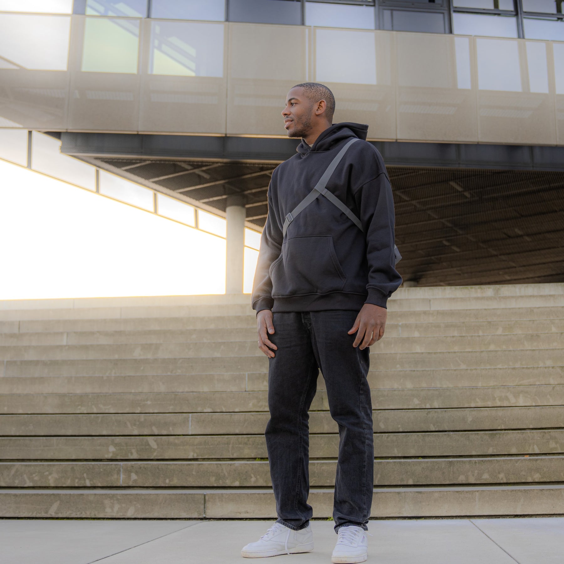 Mann trägt schwarze Umhängetasche 'Dylan' von Johnny Urban, schwarzen Hoodie, Jeans und weiße Sneaker vor modernen Treppen