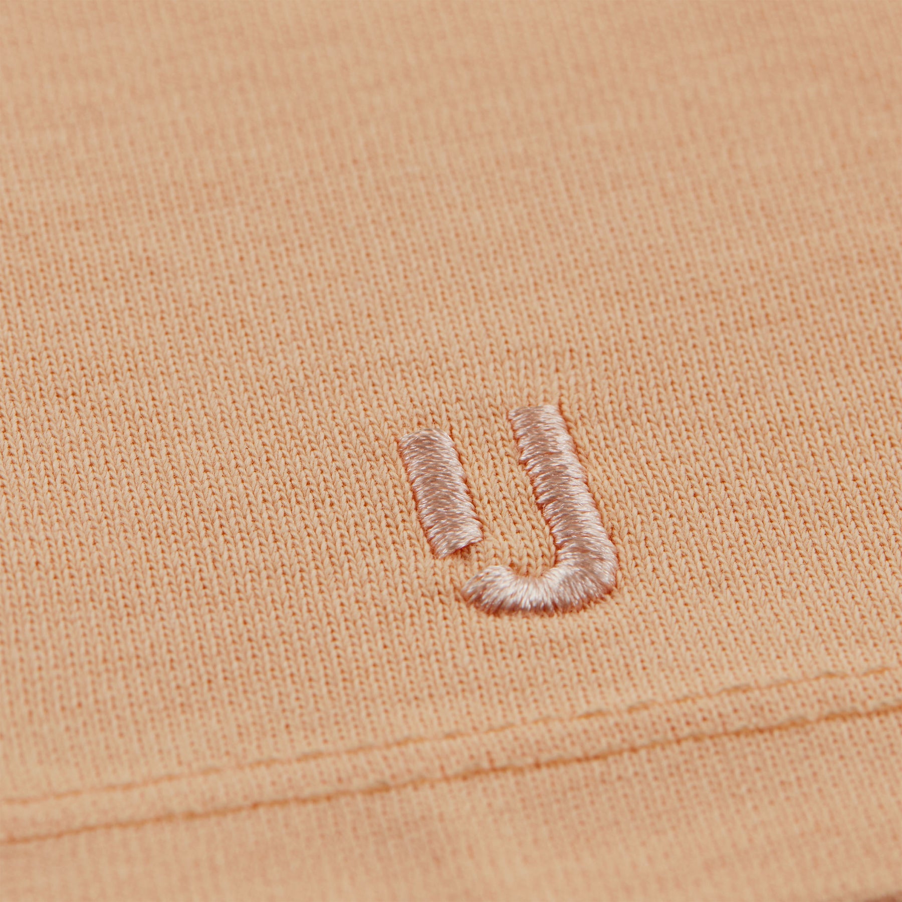 Nahaufnahme der rosa Stickerei des Buchstabens ‚JU‘ auf einem pfirsichfarbenen T-Shirt ‚Sammy‘ von Johnny Urban
