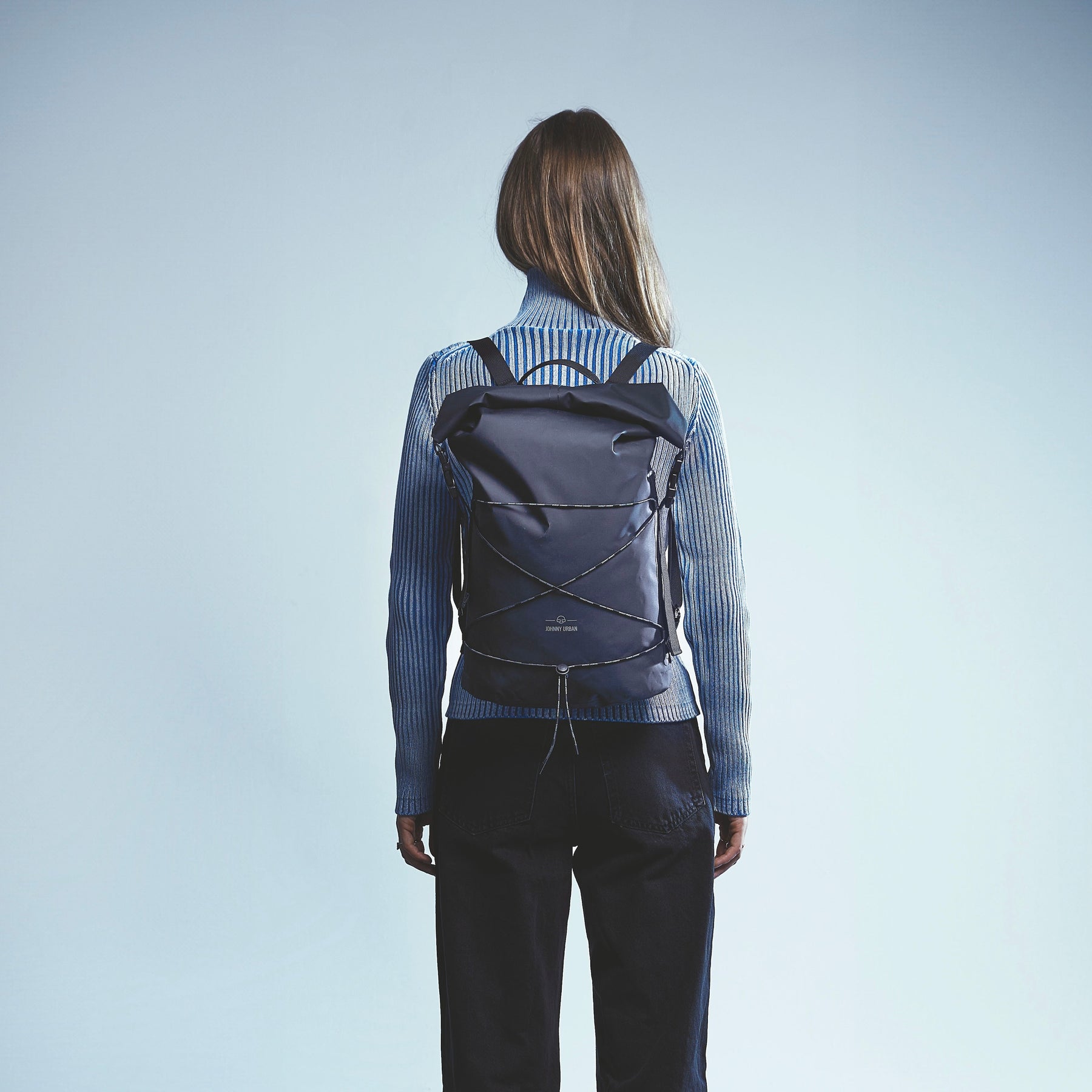Rückansicht einer Frau mit langem braunem Haar, die einen schwarzen rolltop Rucksack 'Myles' von Johnny Urban und einen blauen Rollkragenpullover trägt