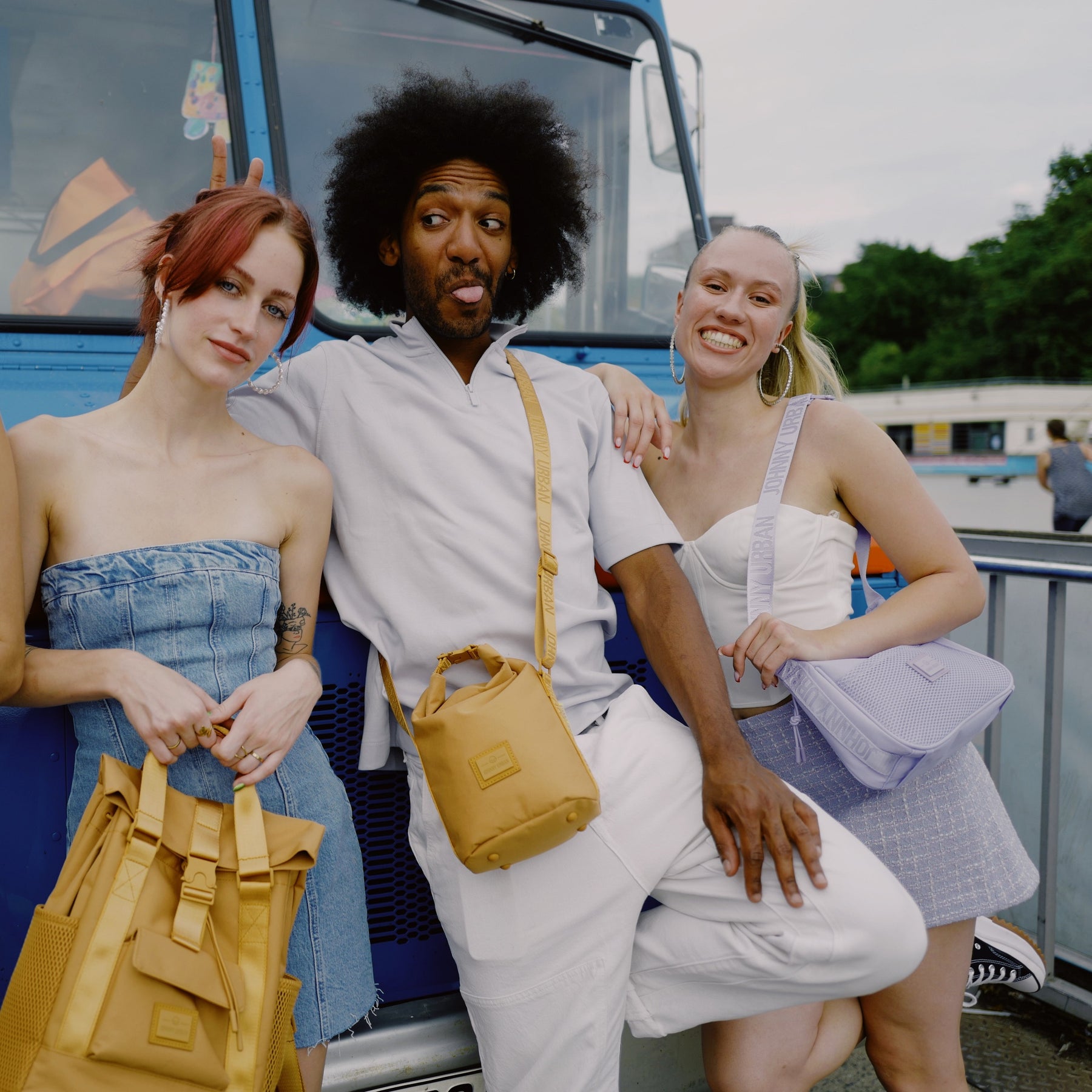 Drei Personen lehnen an blauem Fahrzeug, tragen lässige Kleidung und verschiedene Johnny Urban Taschen, darunter lila Handtasche 'Maddy'