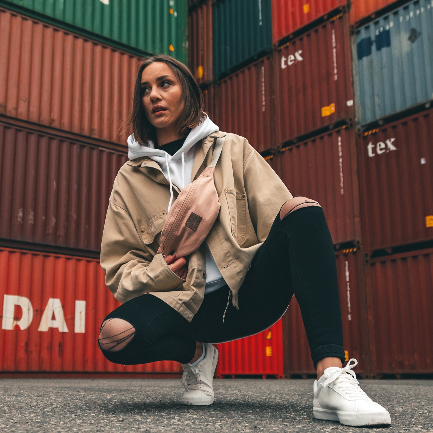 Frau kniet mit rosa Bauchtasche 'Toni', beiger Jacke, weißem Hoodie und zerrissenen schwarzen Jeans vor gestapelten Containern