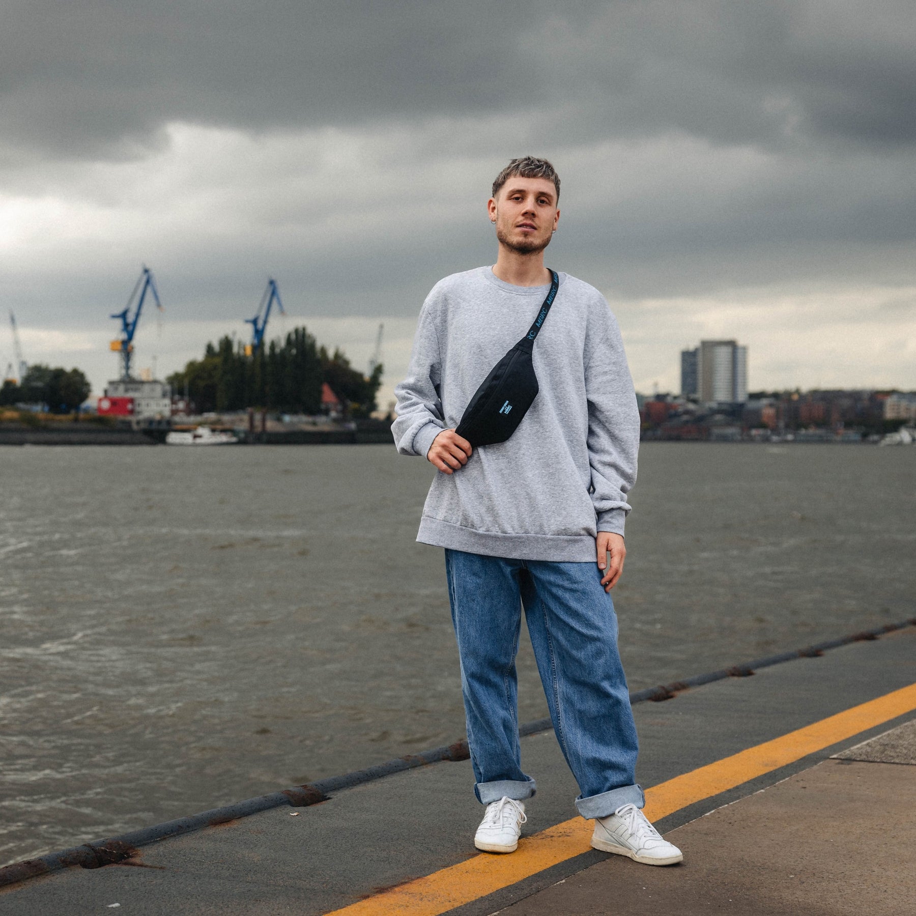 Mann steht am Hafen, trägt eine graue Sweatshirt, blaue Jeans und Johnny Urban Bauchtasche 'Erik' MBRC Edition schwarz quer über die Brust
