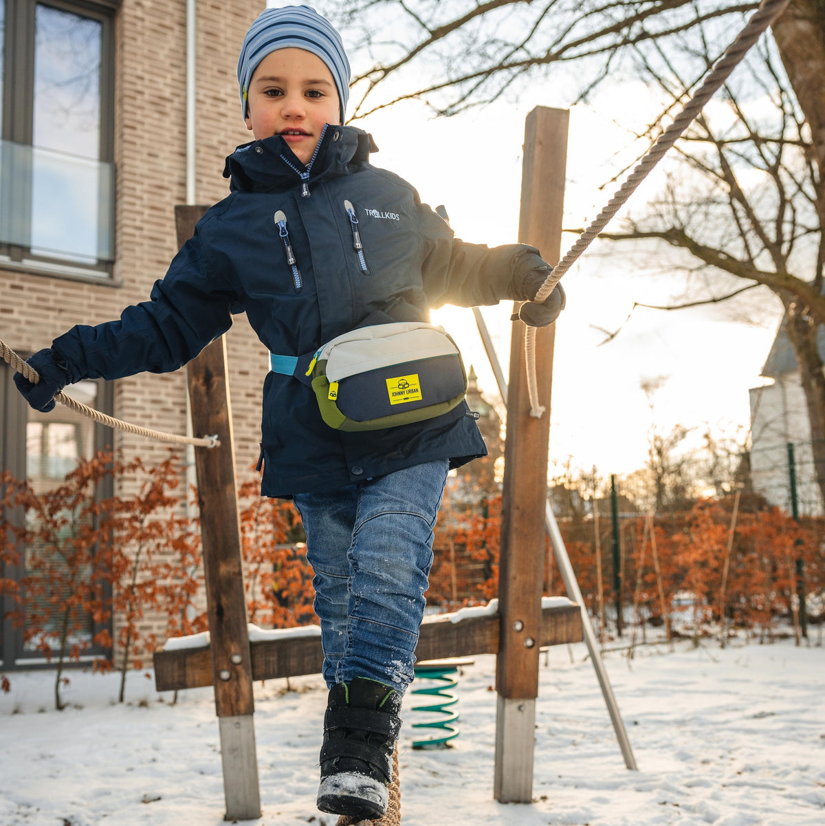 Kind trägt Kinder Bauchtasche 'Felix' in Blau, Weiß und Grün beim Balancieren auf einem Seil im Schnee