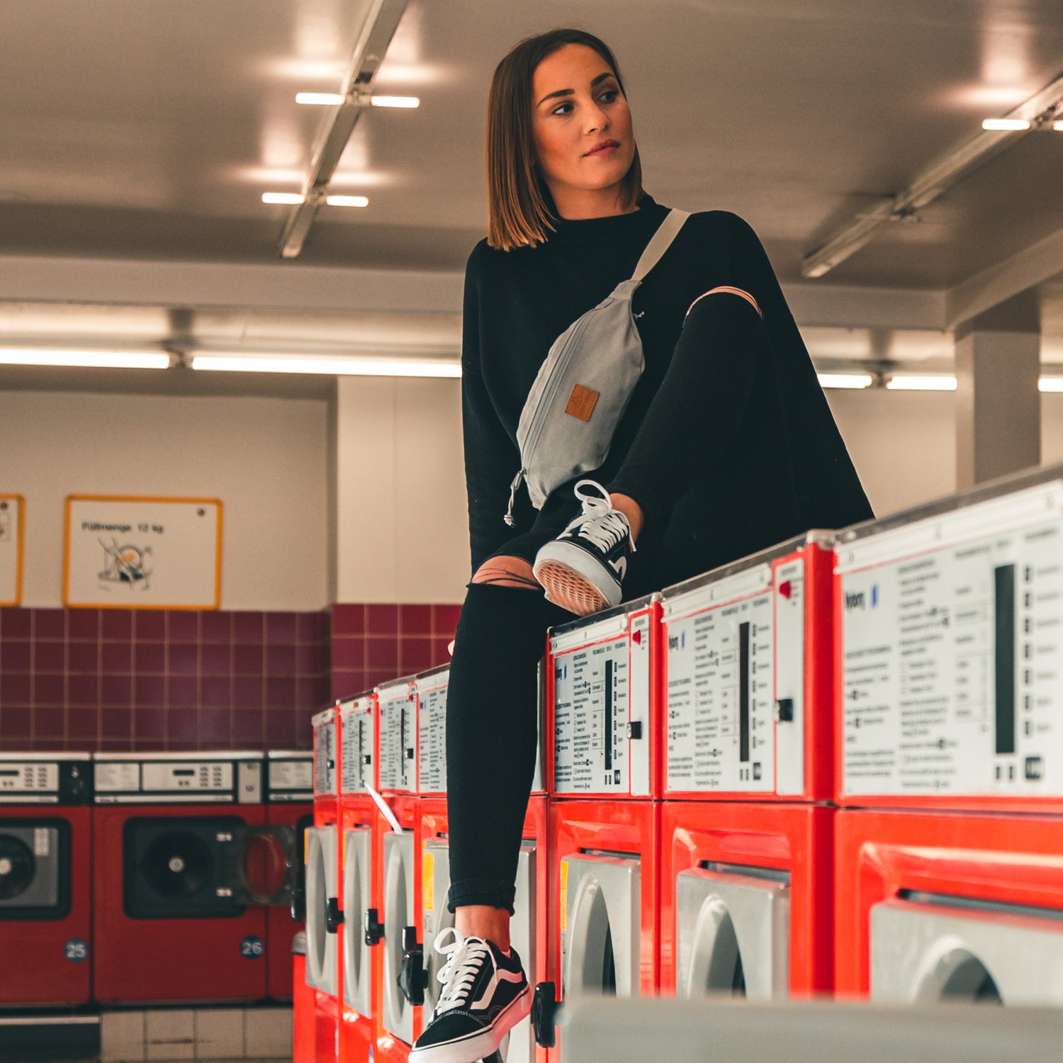 Frau sitzt auf roten Waschmaschinen, trägt schwarze Kleidung und graue Bauchtasche 'Toni' aus Canvas.