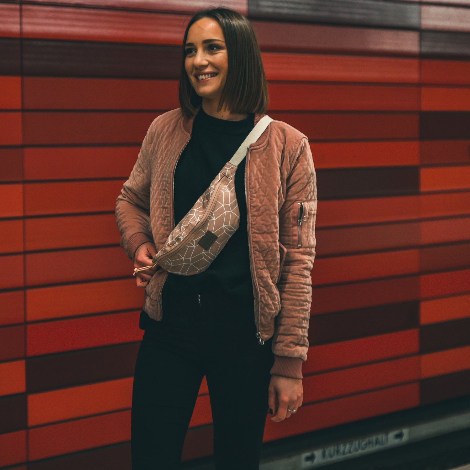 Frau trägt rosafarbene Bauchtasche 'Toni' mit geometrischem Muster diagonall über einer gesteppten Jacke