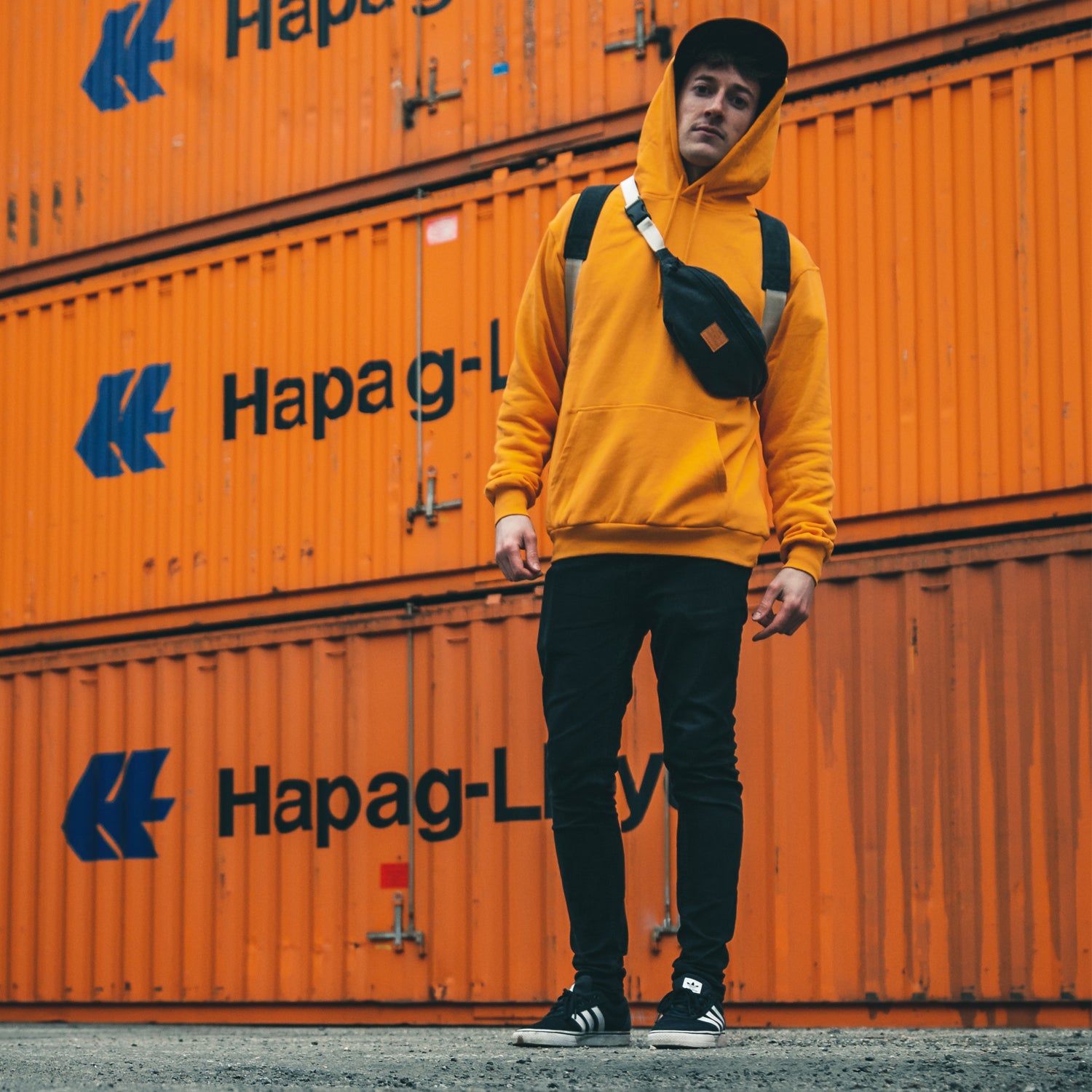 Mann trägt orangenen Hoodie, schwarze Hose, schwarze Cap und anthrazitfarbene Bauchtasche 'Toni' von Johnny Urban vor orangefarbenen Containern