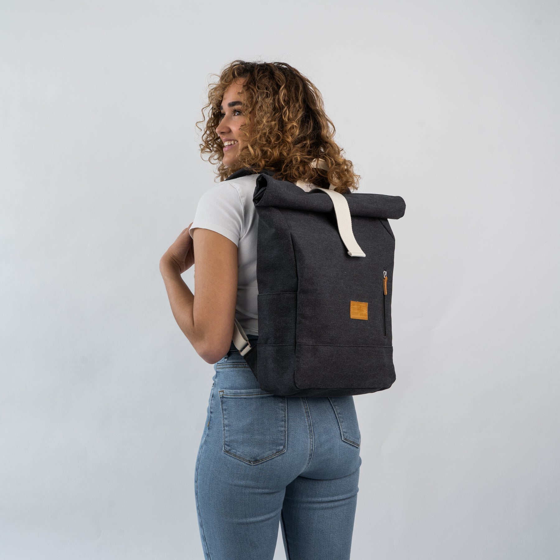 Frau mit lockigen Haaren trägt schwarzen Rolltop Rucksack 'Adam Medium' von Johnny Urban, helles T-Shirt, blaue Jeans