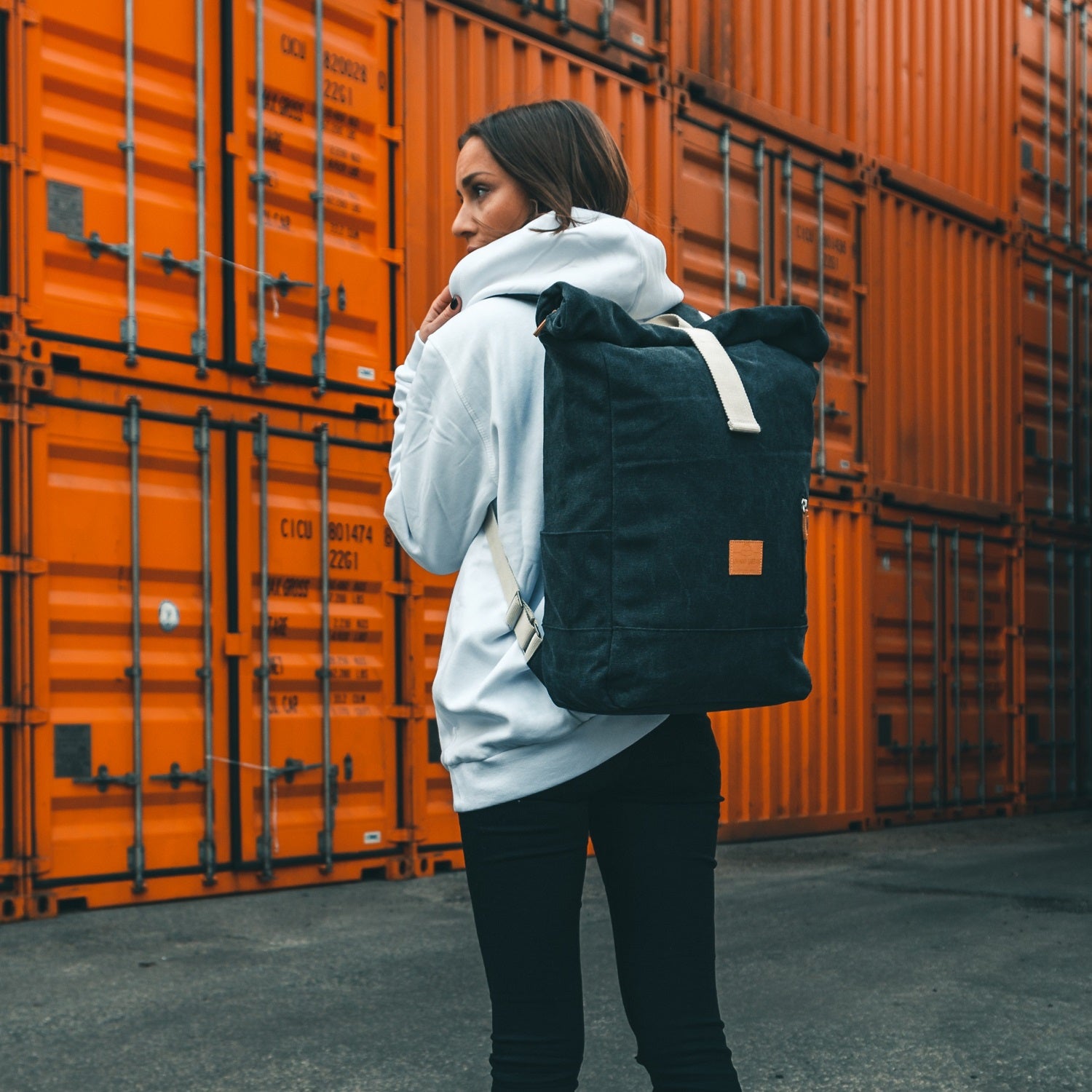 Frau trägt anthrazitfarbenen Rolltop Rucksack 'Adam Large' aus Canvas vor orangefarbenen Containern