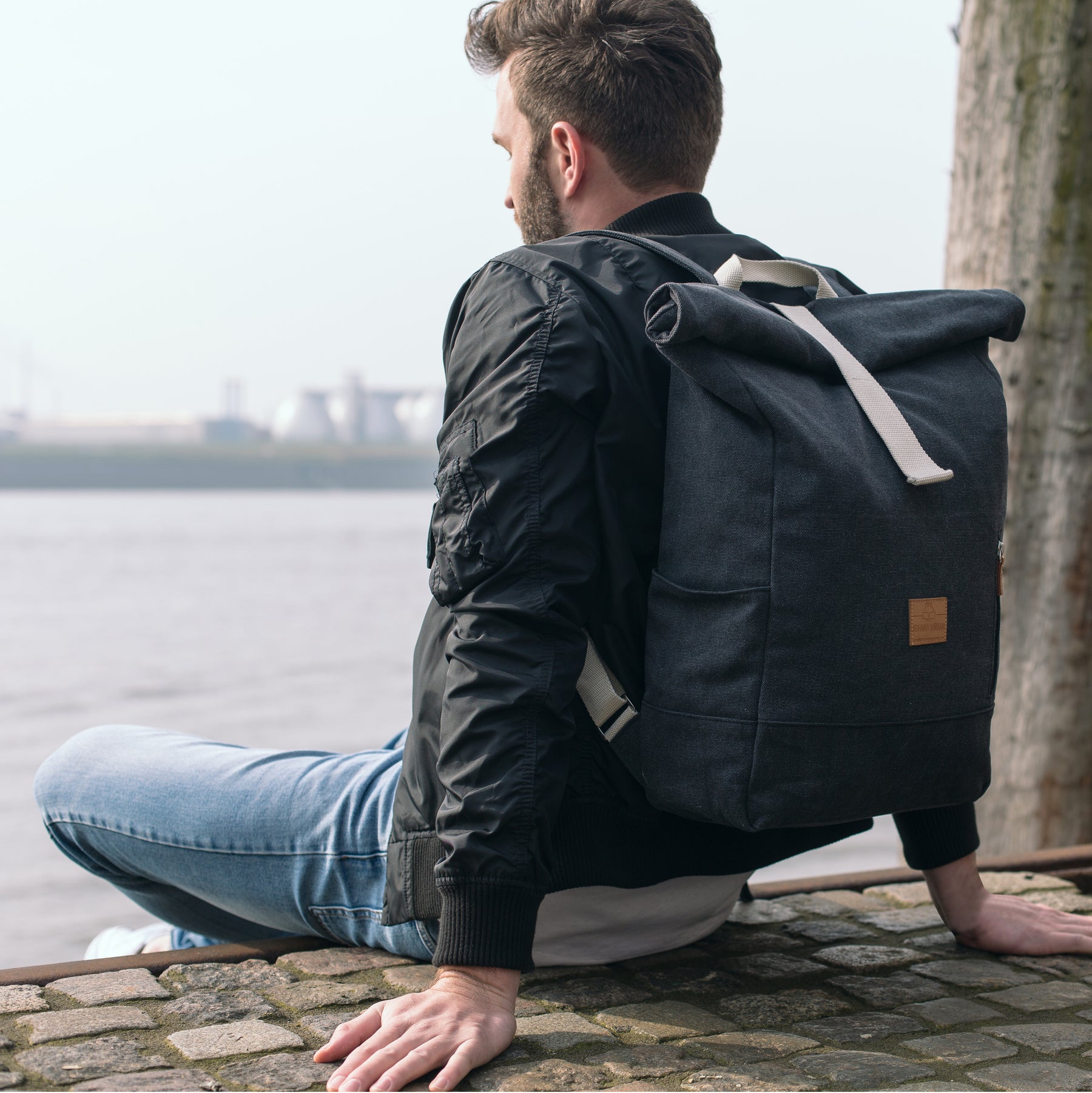 Mann sitzt an Ufer mit anthrazitfarbenem Rolltop Rucksack „Adam Large“ aus Canvas