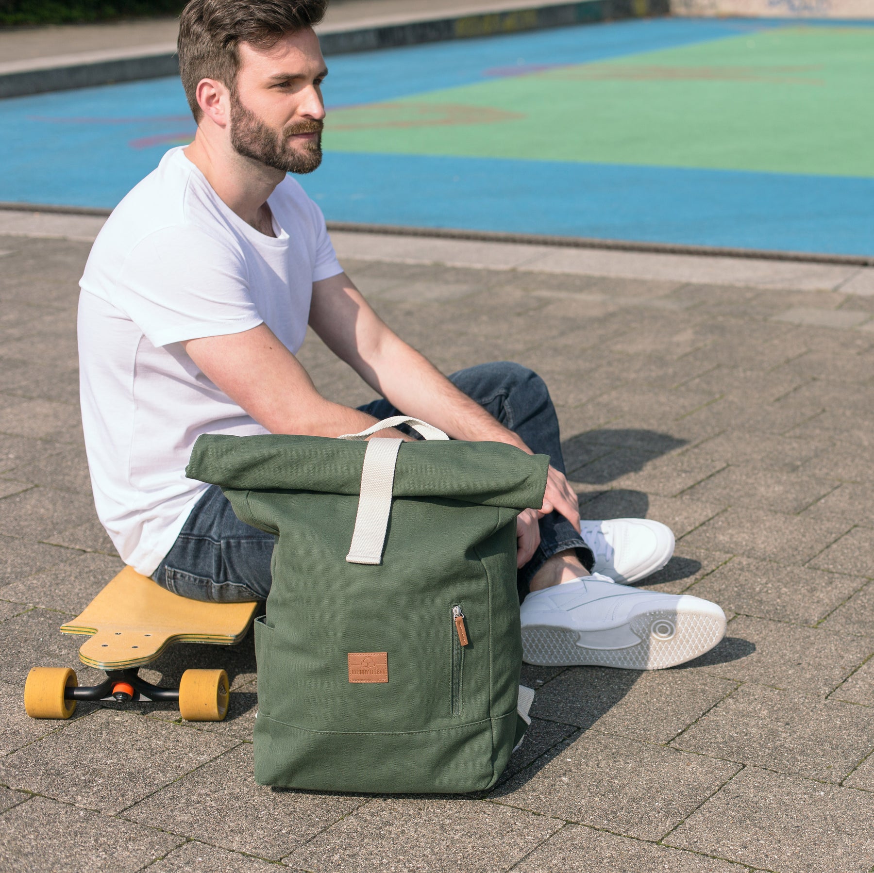 Mann sitzt neben grünem Rolltop-Rucksack 'Adam Large' und gelbem Skateboard auf gepflastertem Boden