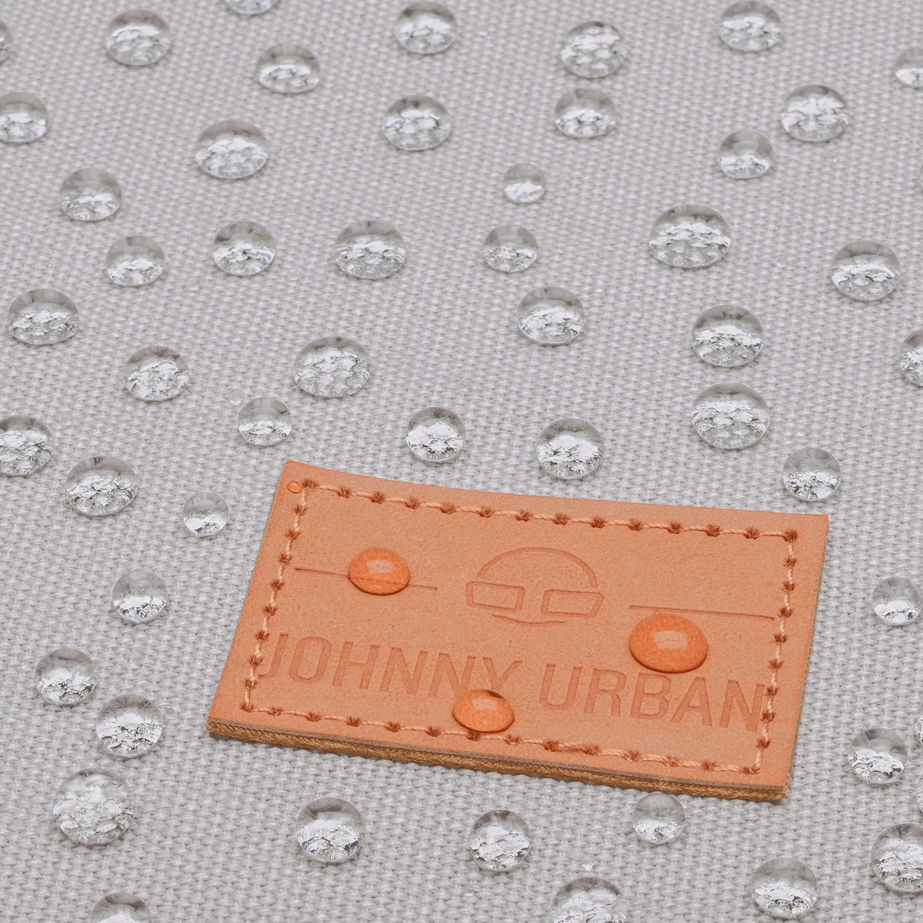 Wasserabweisender grauer Canvasstoff mit aufgenähtem braunen Lederetikett 'Johnny Urban' und Wassertropfen darauf