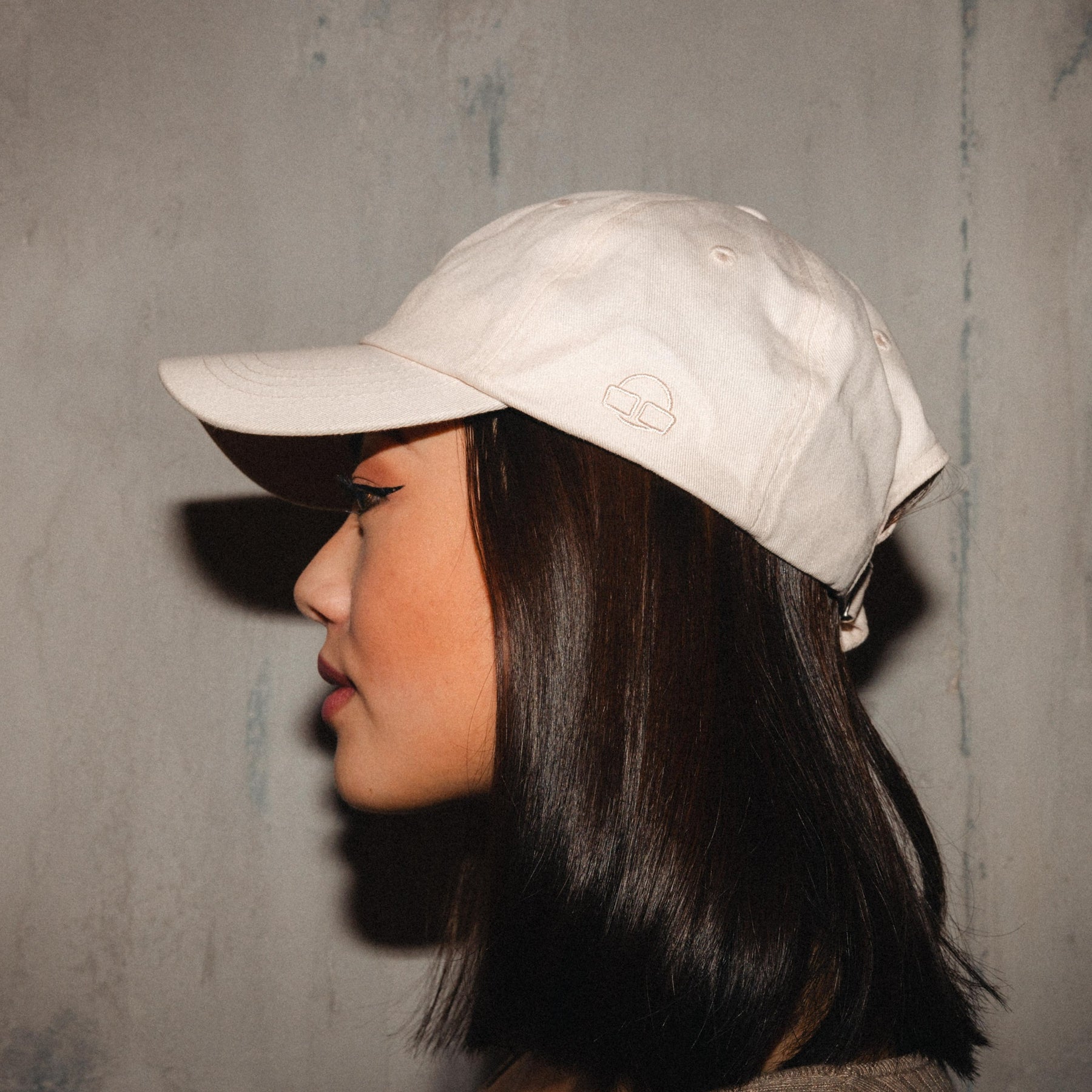 Beige Canvas Cap 'Jen' mit Logo, getragen von Frau im Profil vor grauer Wand
