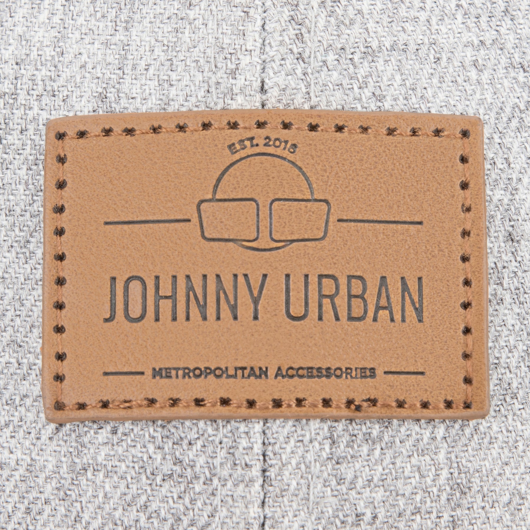 Nahaufnahme eines braunen Lederetiketts mit dem Schriftzug 'JOHNNY URBAN METROPOLITAN ACCESSORIES' auf graubraunem Stoff