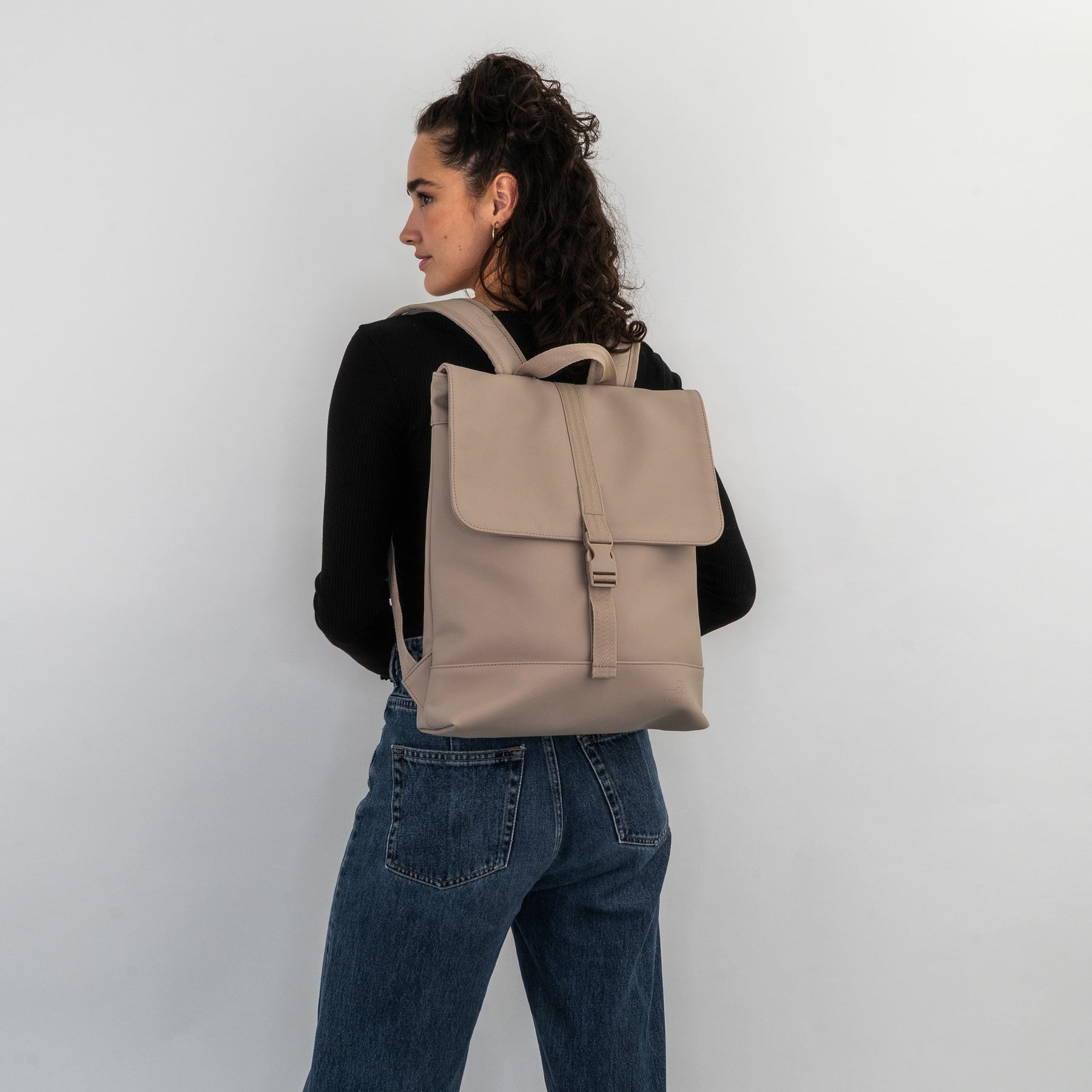 Damen trägt beigefarbenen Rucksack 'Ruby' mit Klappe und Schnalle, schwarze Oberteile, blaue Jeans