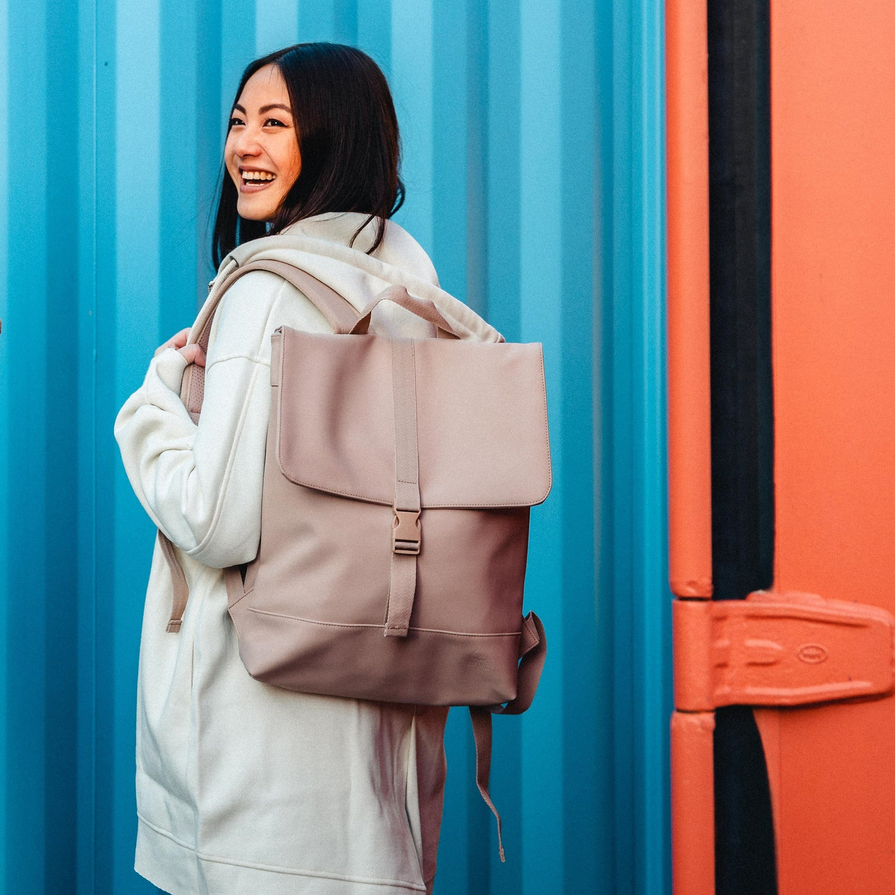 Frau trägt den rosa Damen-Rucksack 'Ruby' mit Klappe und Schnalle vor blau-orange Hintergrund