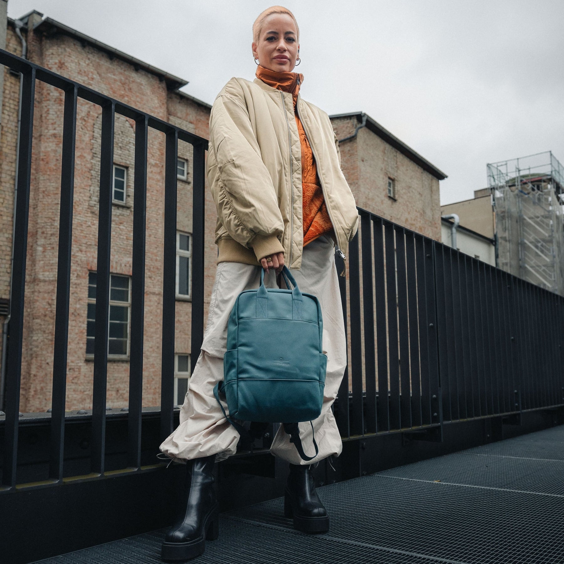 Frau in beigem Bomberjacke, oranger Jacke und weiten hellen Hosen hält petrolgrünen Rucksack 'Hailey'