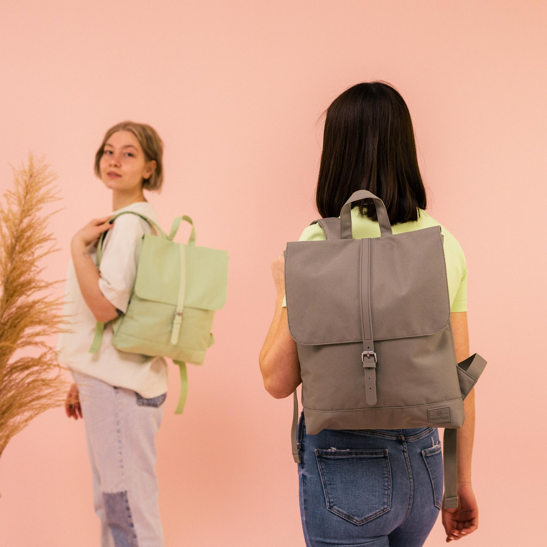 Zwei Frauen mit nachhaltigen Damenrucksäcken 'Mia' von Johnny Urban, eine braun, eine hellgrün, vor rosa Hintergrund
