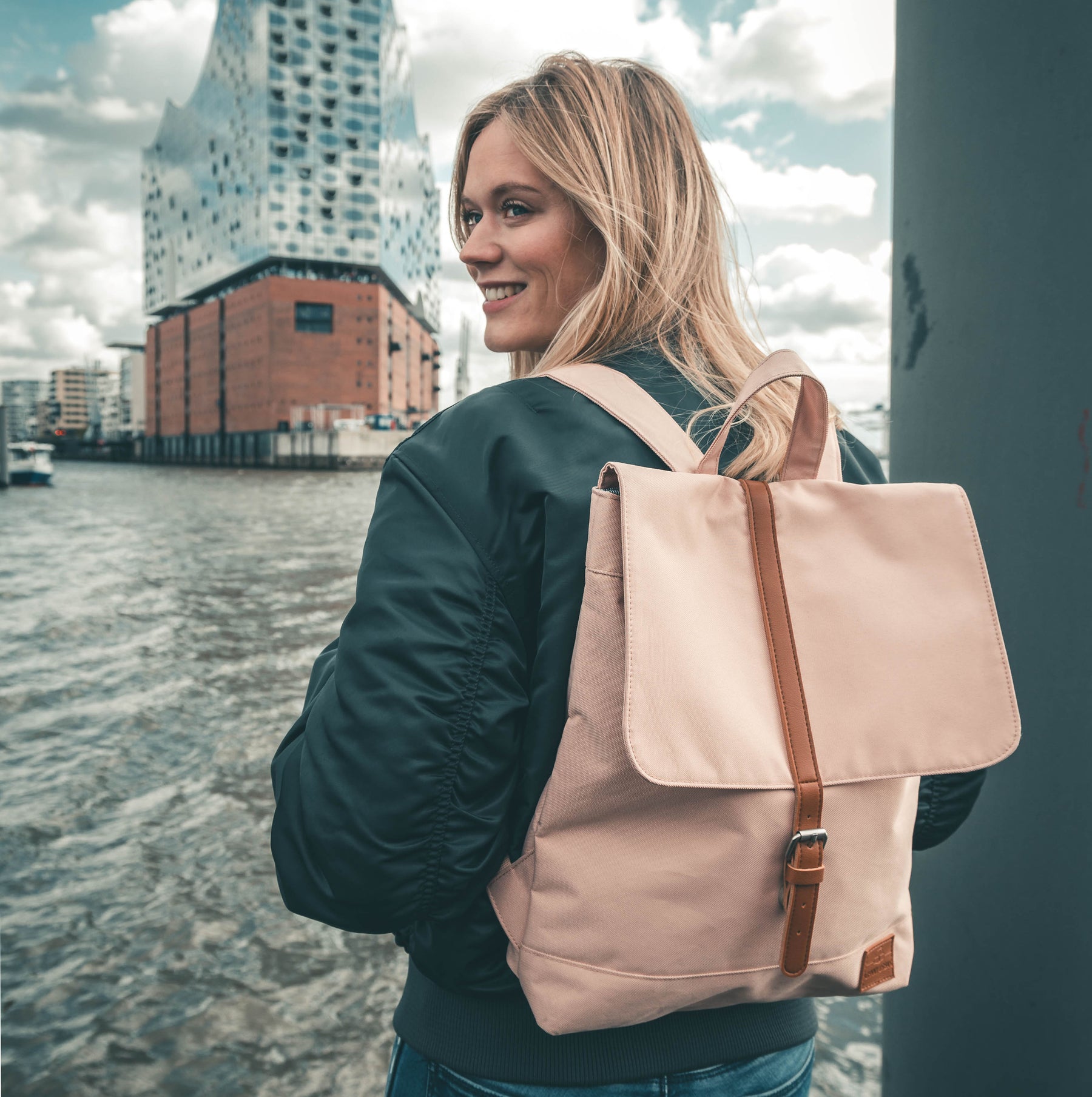 Frau mit schwarzer Jacke trägt rosa Damenrucksack 'Mia' von Johnny Urban an einem Hafen mit modernem Gebäude