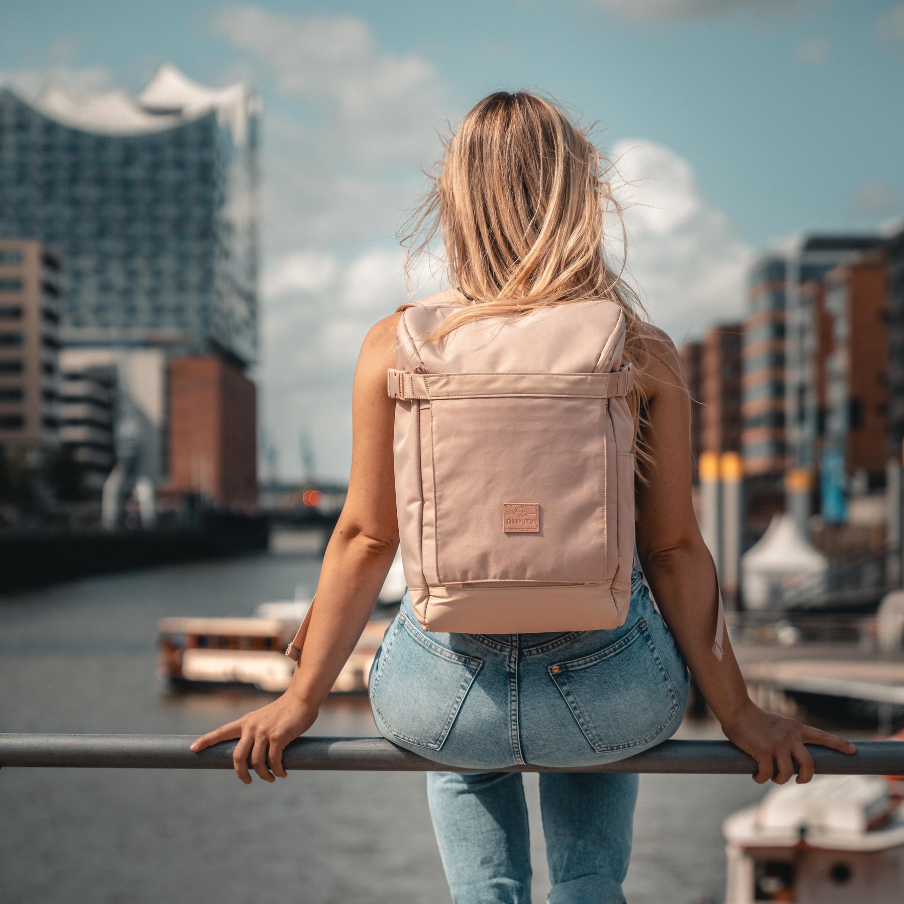 Frau mit rosa Rucksack 'Luca' sitzt an Geländer am Wasser in urbaner Umgebung