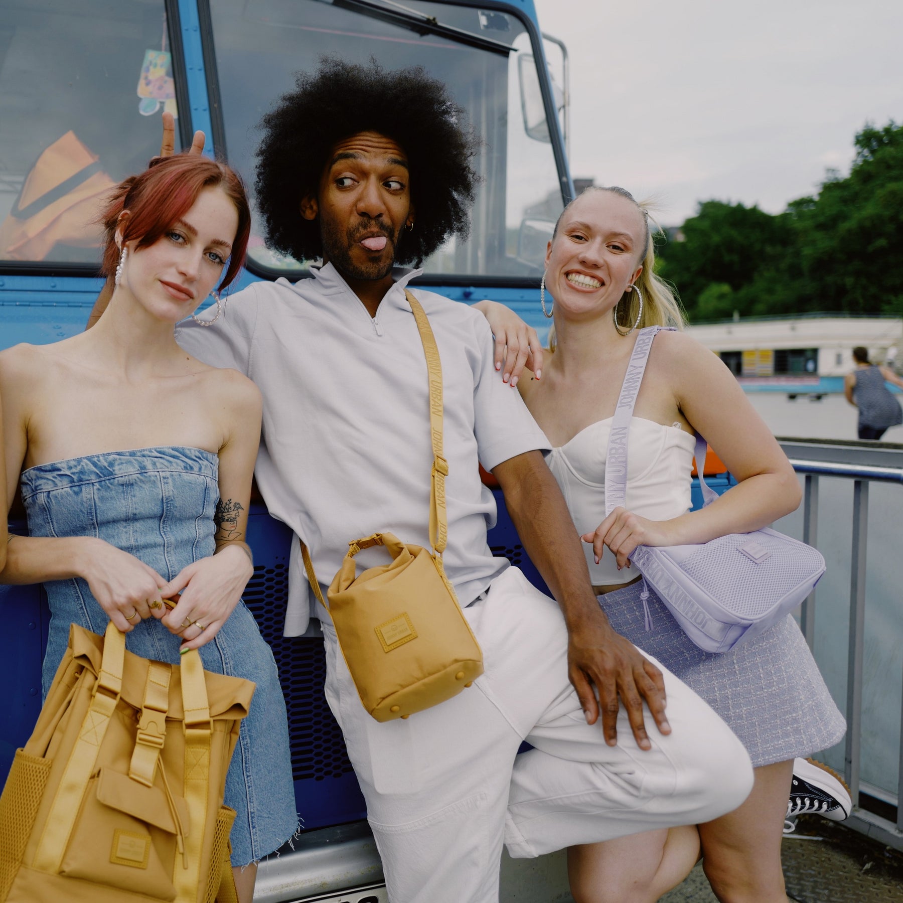 Drei junge Personen vor blauem Fahrzeug, Mann trägt caramelbraune Umhängetasche 'Rue' von Johnny Urban