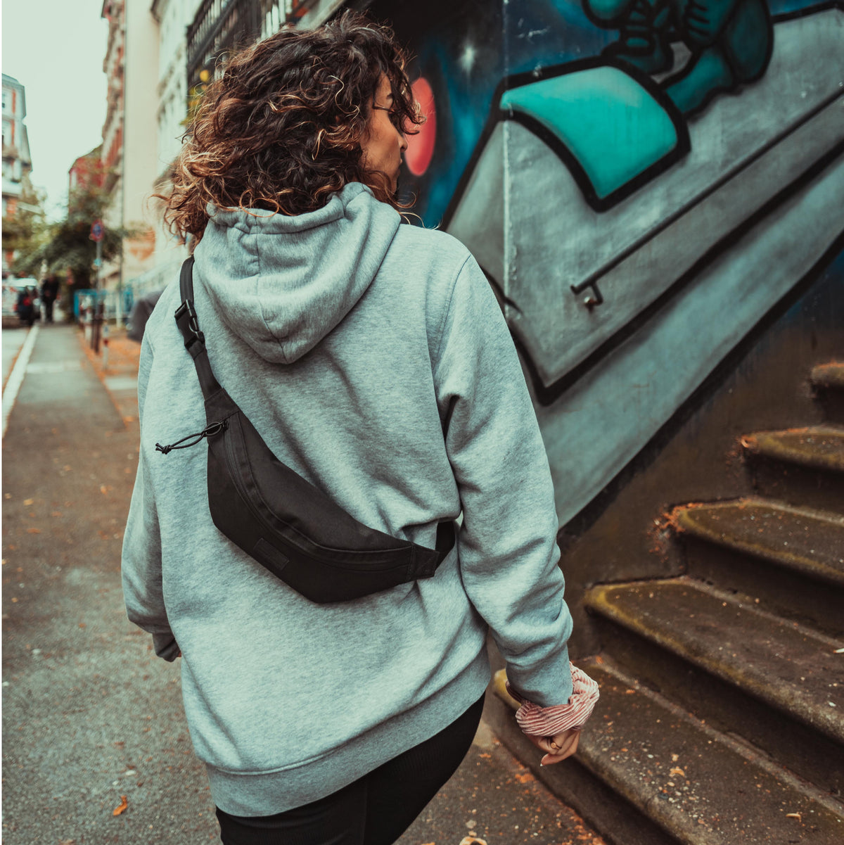 Frau mit lockigem Haar, grauem Hoodie und schwarzer Bauchtasche ‚Erik Small‘ trägt Tasche über Rücken in urbaner Umgebung