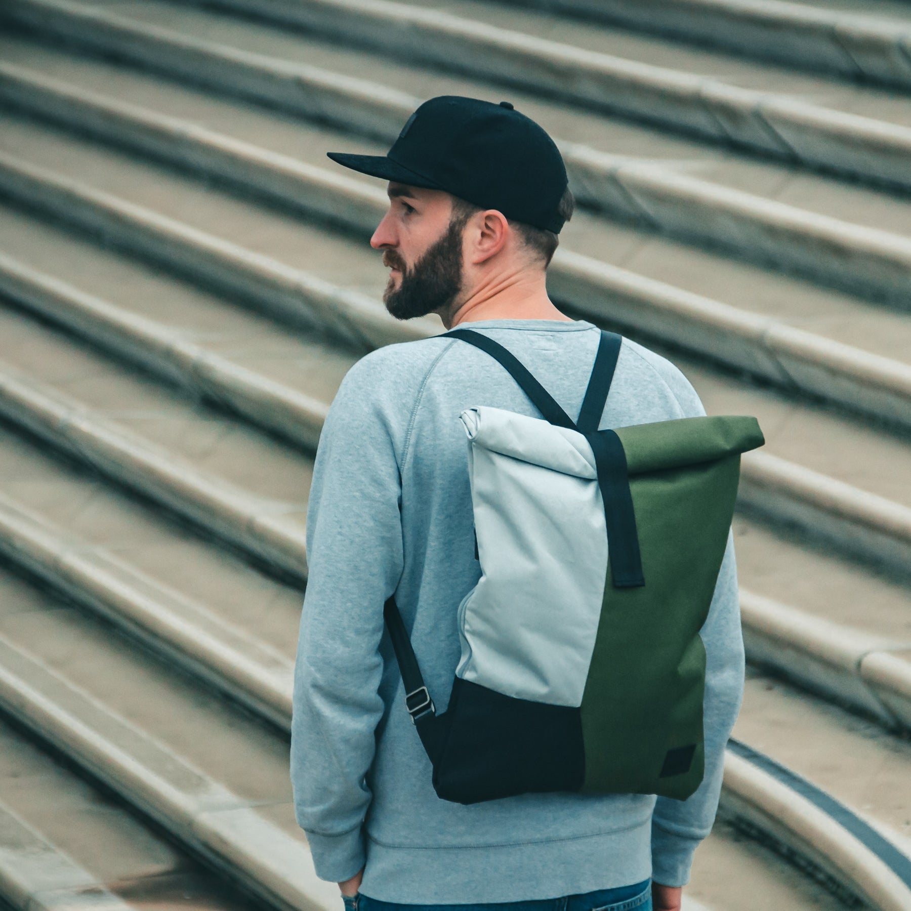 Mann mit schwarzer Cap 'Dean', grauem Pullover und zweifarbigen Rucksack vor einer Treppe