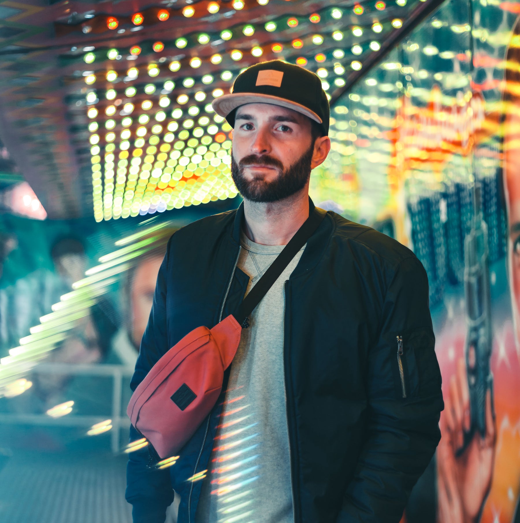 Mann mit schwarzer 'Dean'-Cap, roter Gürteltasche und schwarzer Jacke vor leuchtender urbaner Kulisse