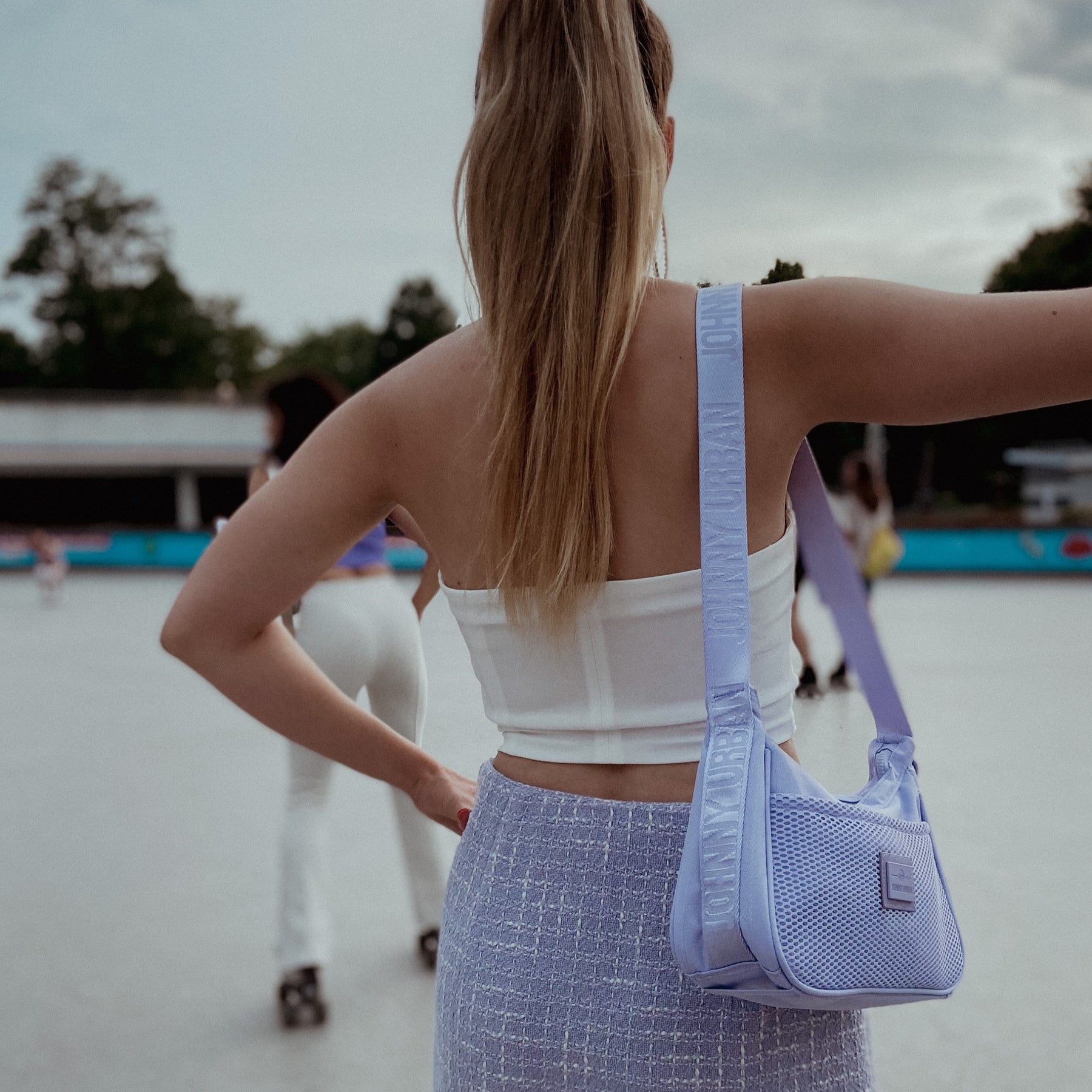 Rückansicht einer Frau mit Pferdeschwanz, die eine lila Johnny Urban Handtasche 'Maddy' trägt, mit weißem Top und lilafarbenem Rock