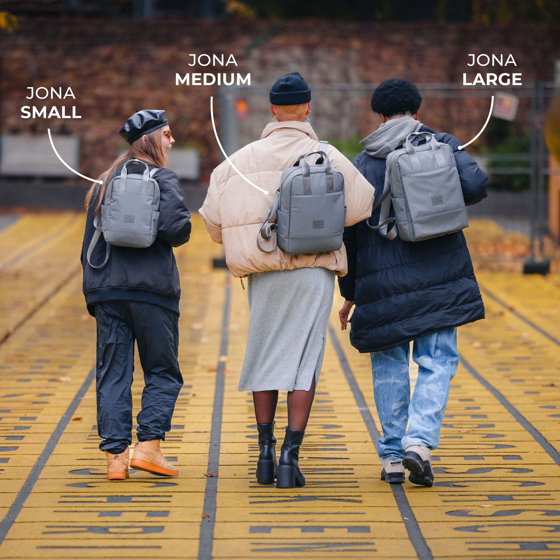 Drei Personen gehen auf gelb markiertem Boden, tragen graue Jona Rücksäcke in den Größen Small, Medium und Large auf dem Rücken