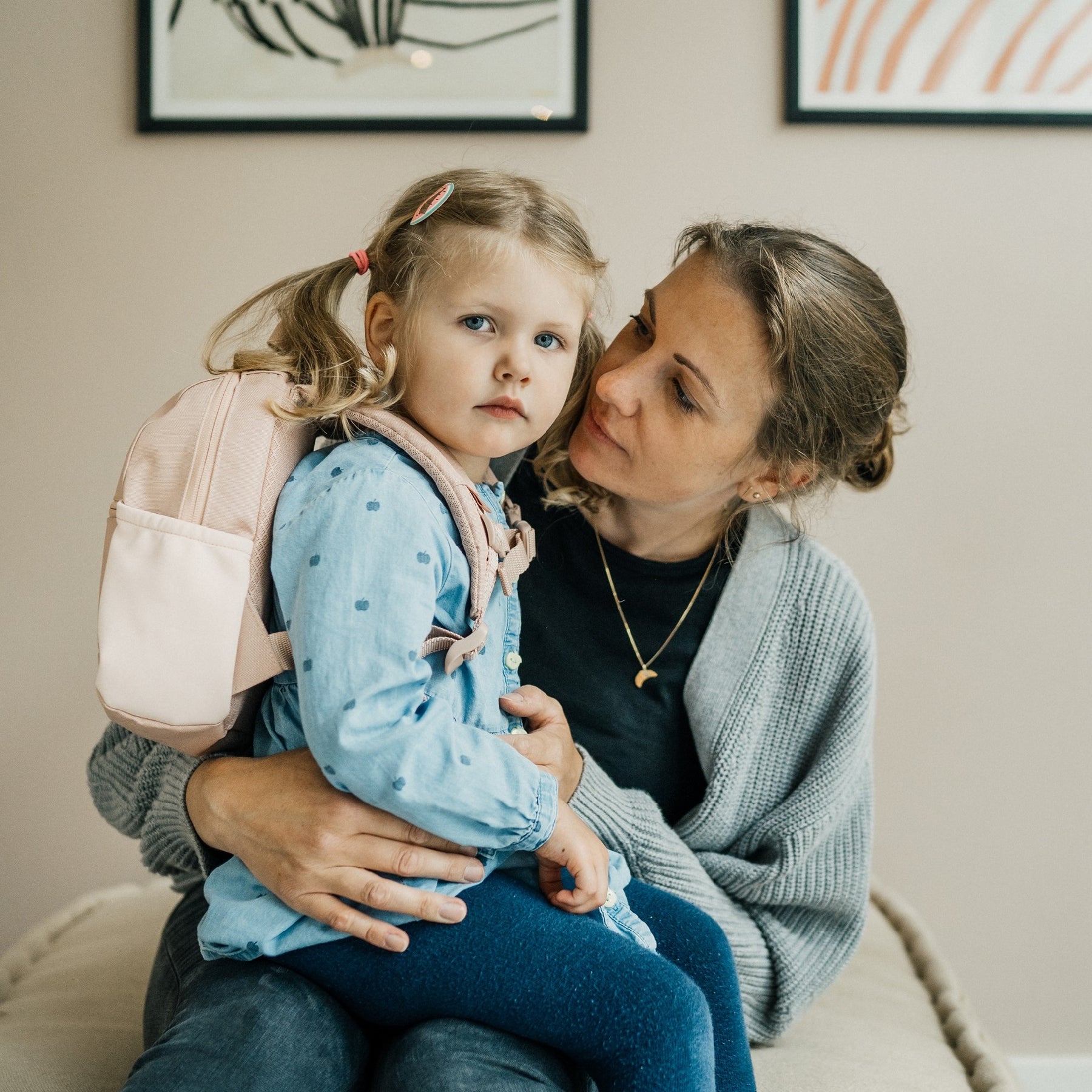 Kleines Mädchen mit rosa Kinderrucksack 'Leo' sitzt auf dem Schoß einer Frau in grauem Strickcardigan