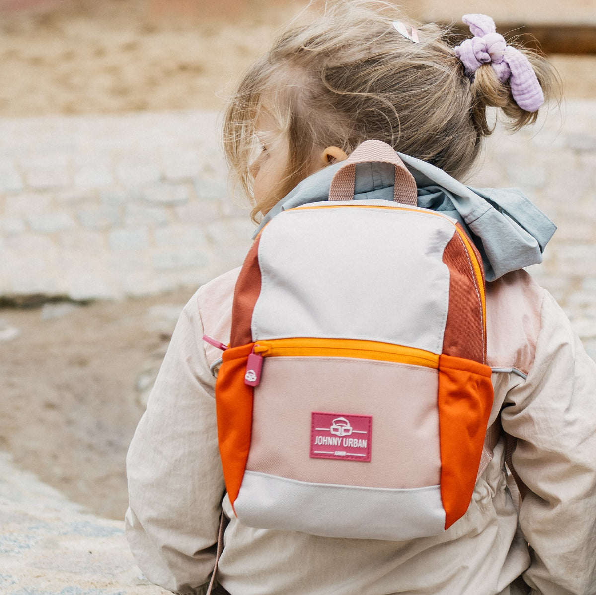 Kinderrucksack 'Leo' in Beige, Orange und Braun getragen von einem Mädchen mit hellbraunem Haar und lilafarbenem Haargummi