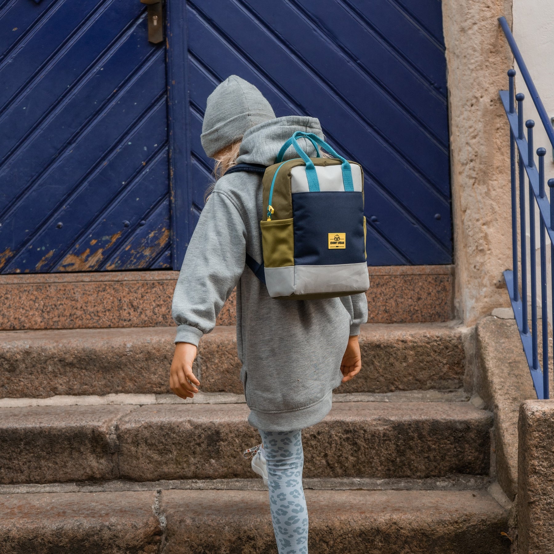 Kinderrucksack 'Linus', rechteckig in Blau, Grün und Grau, auf dem Rücken eines Kindes mit grauer Kapuzenjacke und Mütze