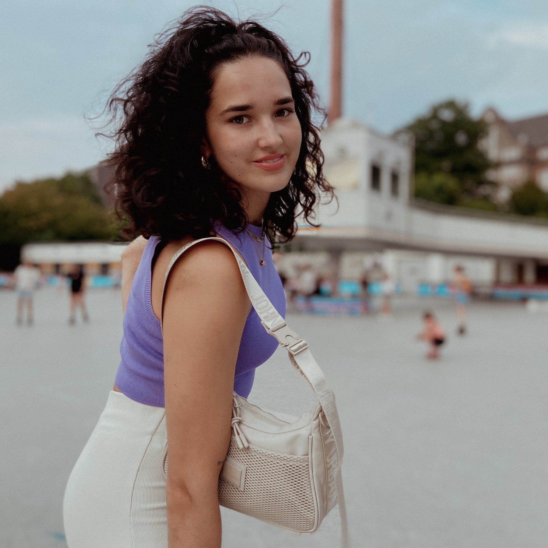 Frau trägt lilafarbenes Top und weiße Hose, beiges Handtasche Modell 'Maddy' über der Schulter