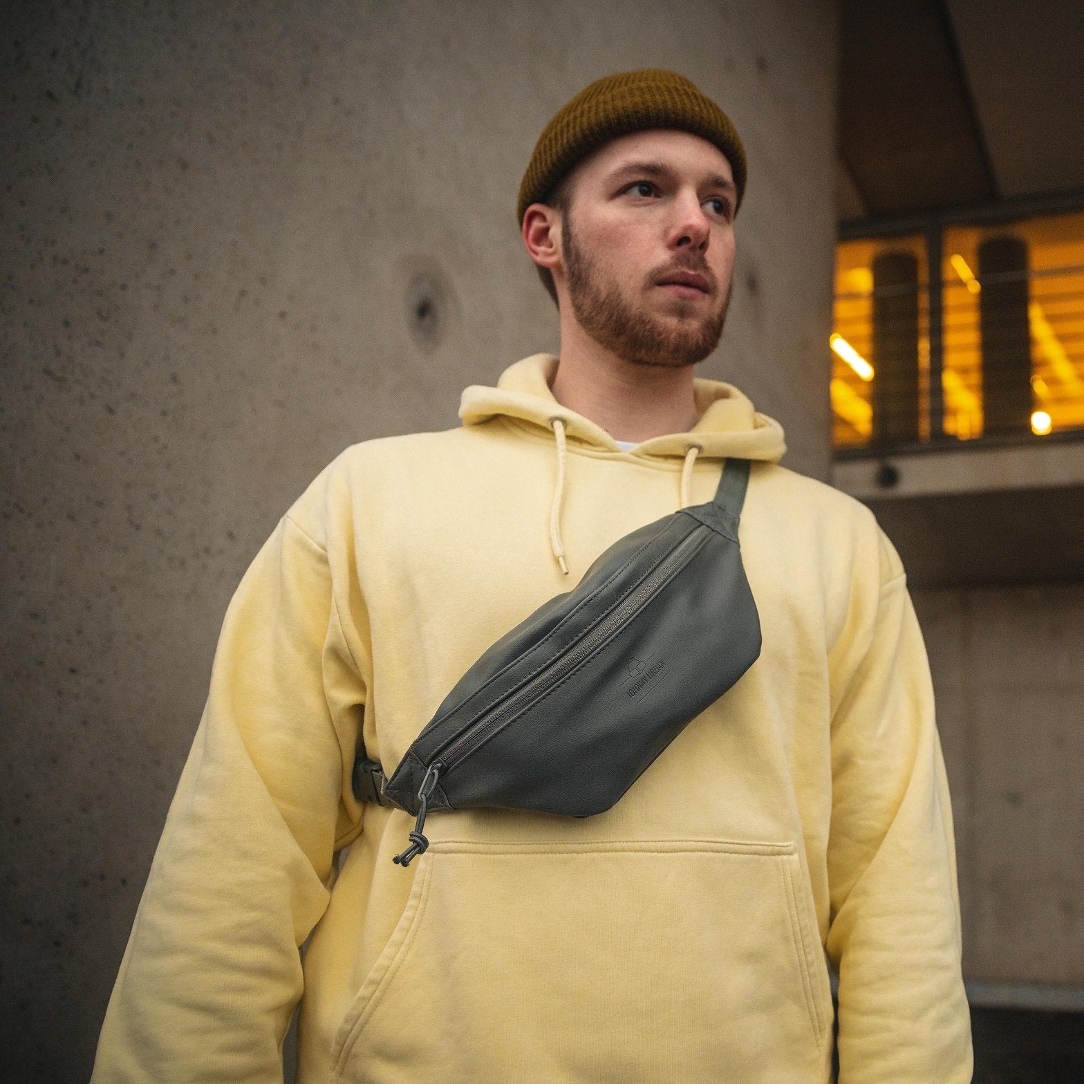 Mann mit gelbem Hoodie und brauner Mütze trägt graue Kunstleder-Bauchtasche "Toni" von Johnny Urban quer über die Brust