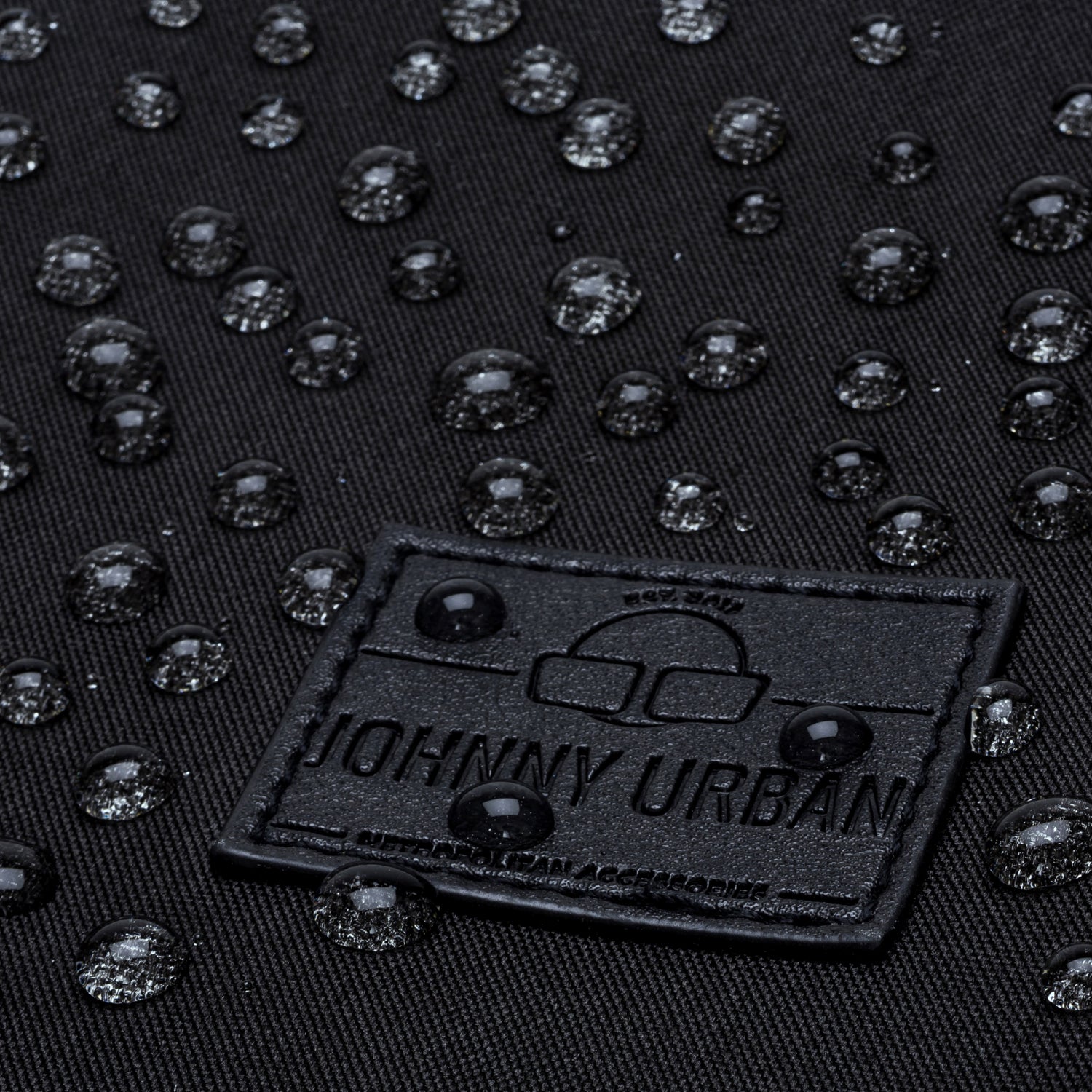 Nahe Aufnahme des schwarzen Labels des Johnny Urban Rucksacks Jona Large mit Wassertropfen auf schwarzem Stoff