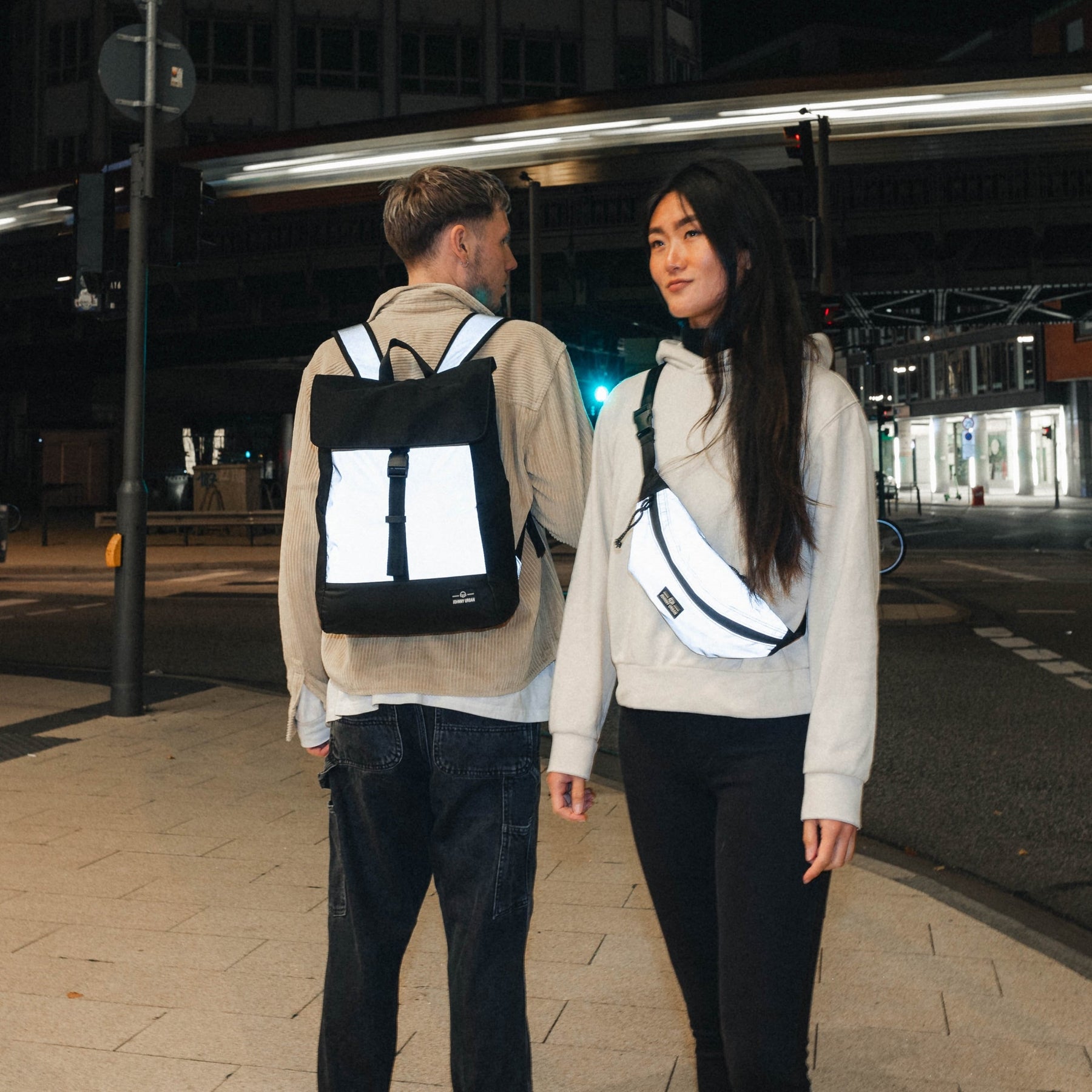 Mann trägt reflektierenden schwarzen Rucksack 'Mika', Frau trägt reflektierende Umhängetasche bei Nacht in Stadt