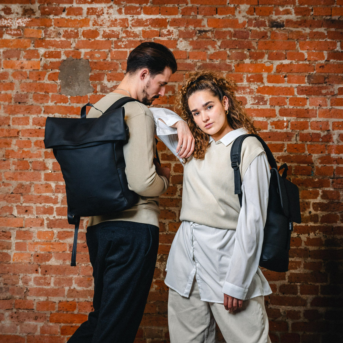 Mann und Frau tragen schwarzen Rolltop Rucksack 'Henry' von Johnny Urban vor Ziegelsteinwand