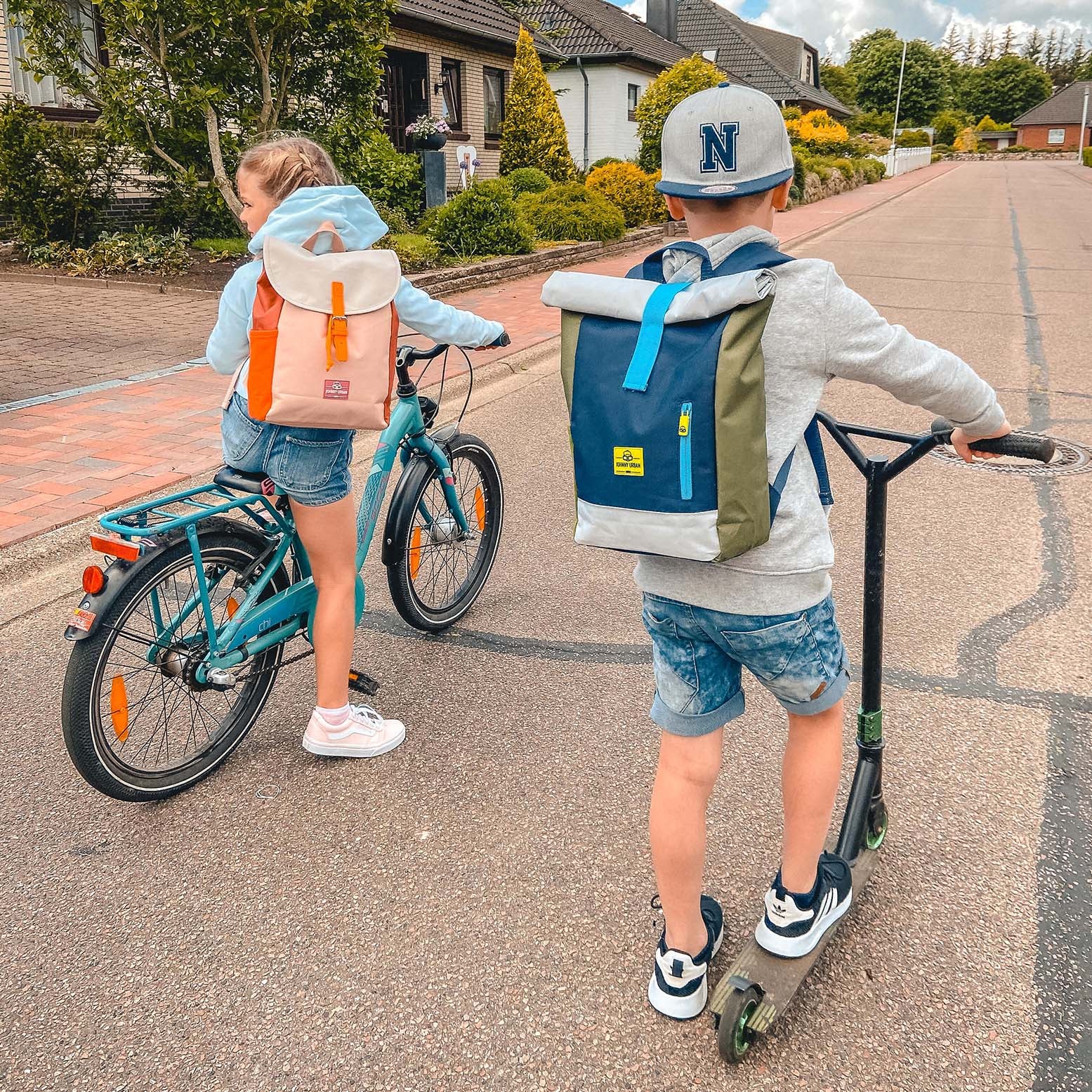 Zwei Kinder mit Rolltop-Kinderrucksäcken Aaron, ein Mädchen auf einem Fahrrad mit rosa-orangenem Rucksack, ein Junge auf einem Roller mit blau-grünem Rucksack