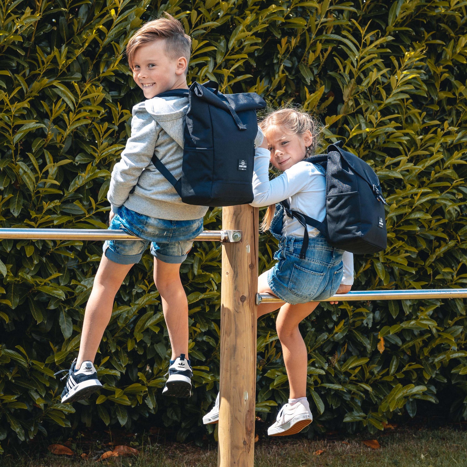 Zwei Kinder mit schwarzen Rolltop-Kinderrucksäcken ‚Aaron‘ klettern auf ein Holzgeländer im Freien