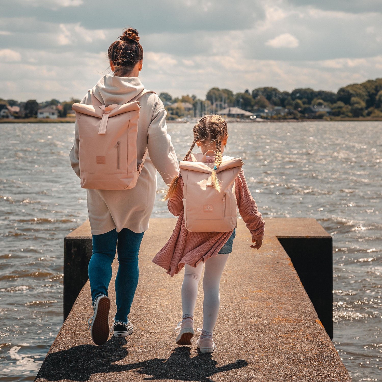 Mutter und Kind laufen auf einem Steg am Wasser, tragen rosa Rolltop Kinderrucksäcke von Johnny Urban