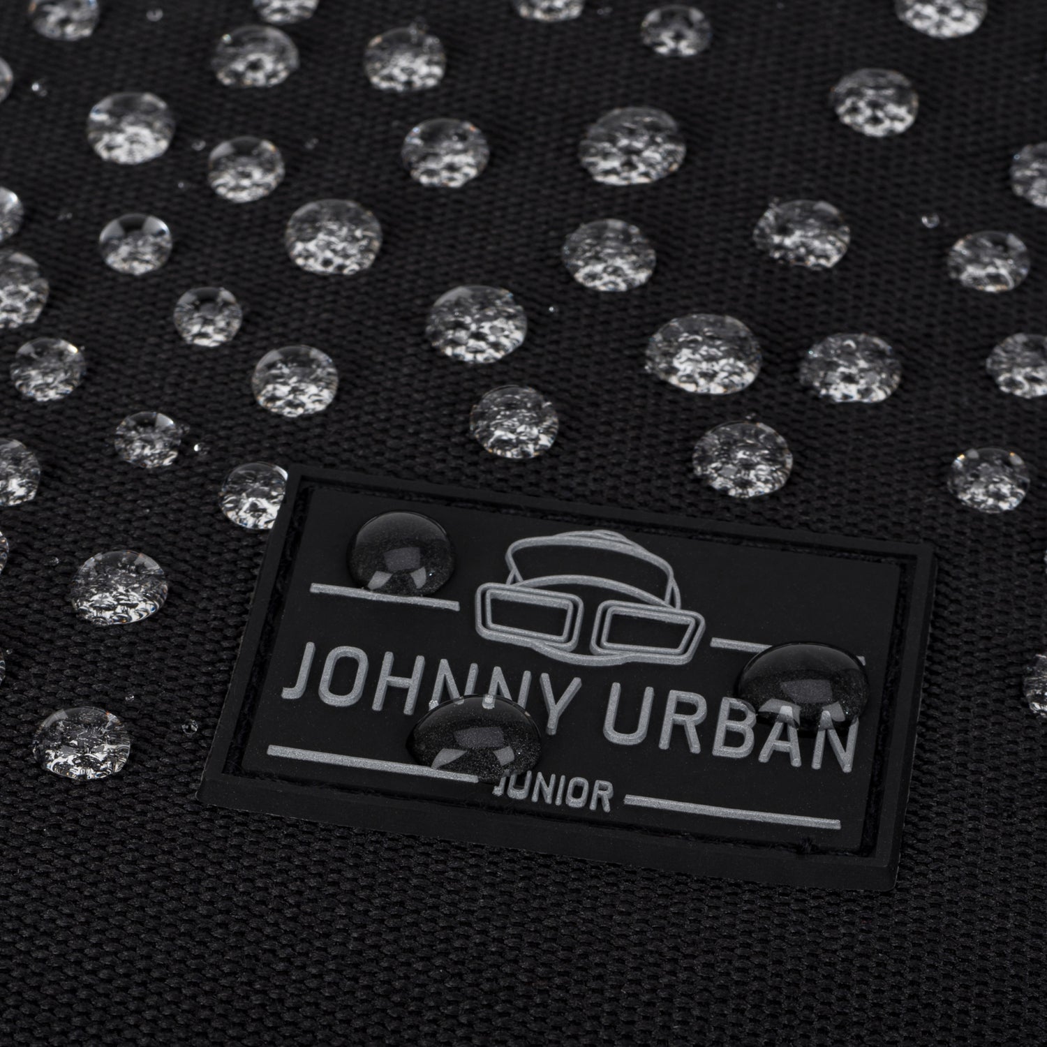 Nahaufnahme eines schwarzen wasserdichten Rolltop Kinderrucksacks 'Aaron' mit Johnny Urban Junior Logo und Wasserperlen