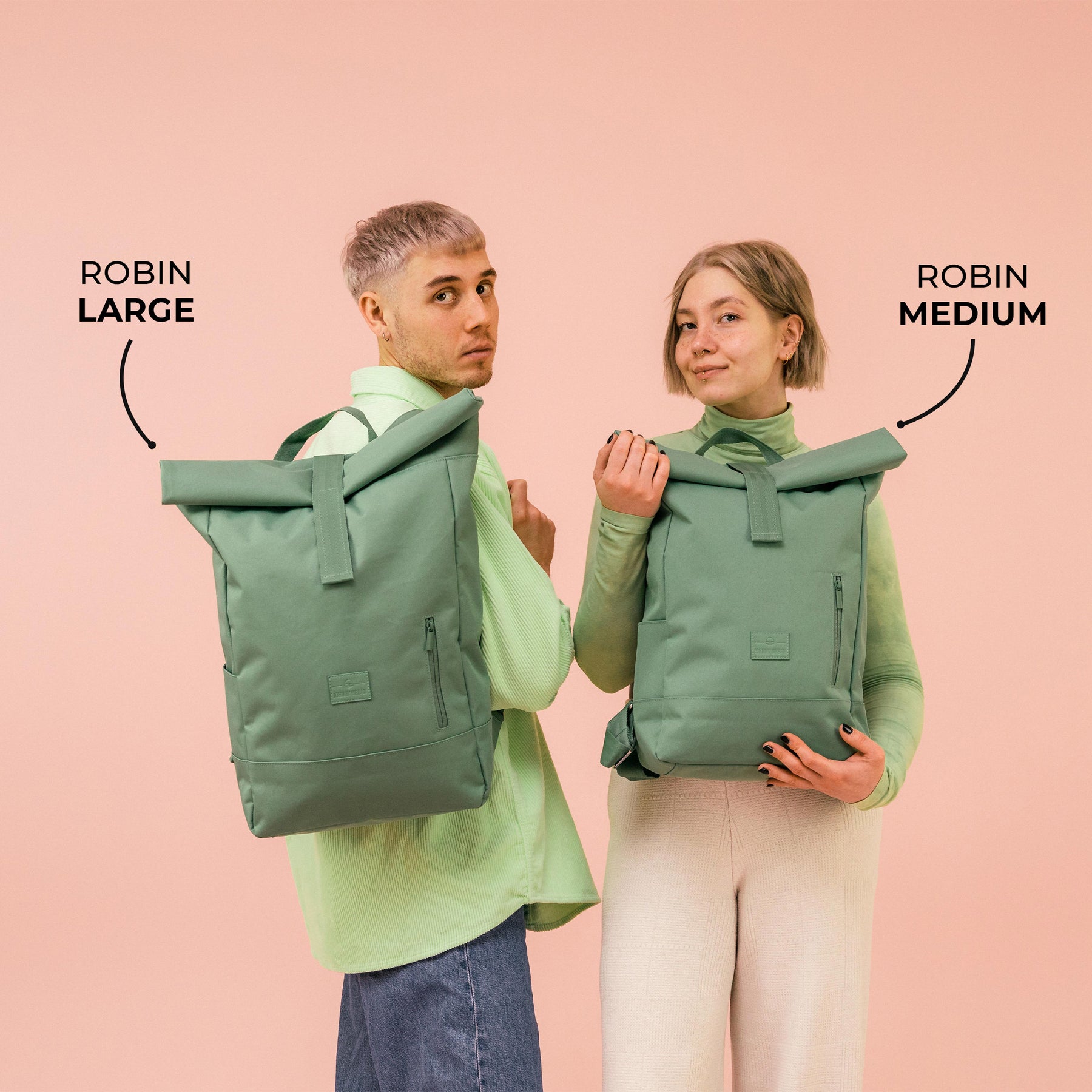 Zwei Personen zeigen Rolltop-Rucksäcke 'Robin Large' und 'Robin Medium' in Salbeigrün vor rosa Hintergrund