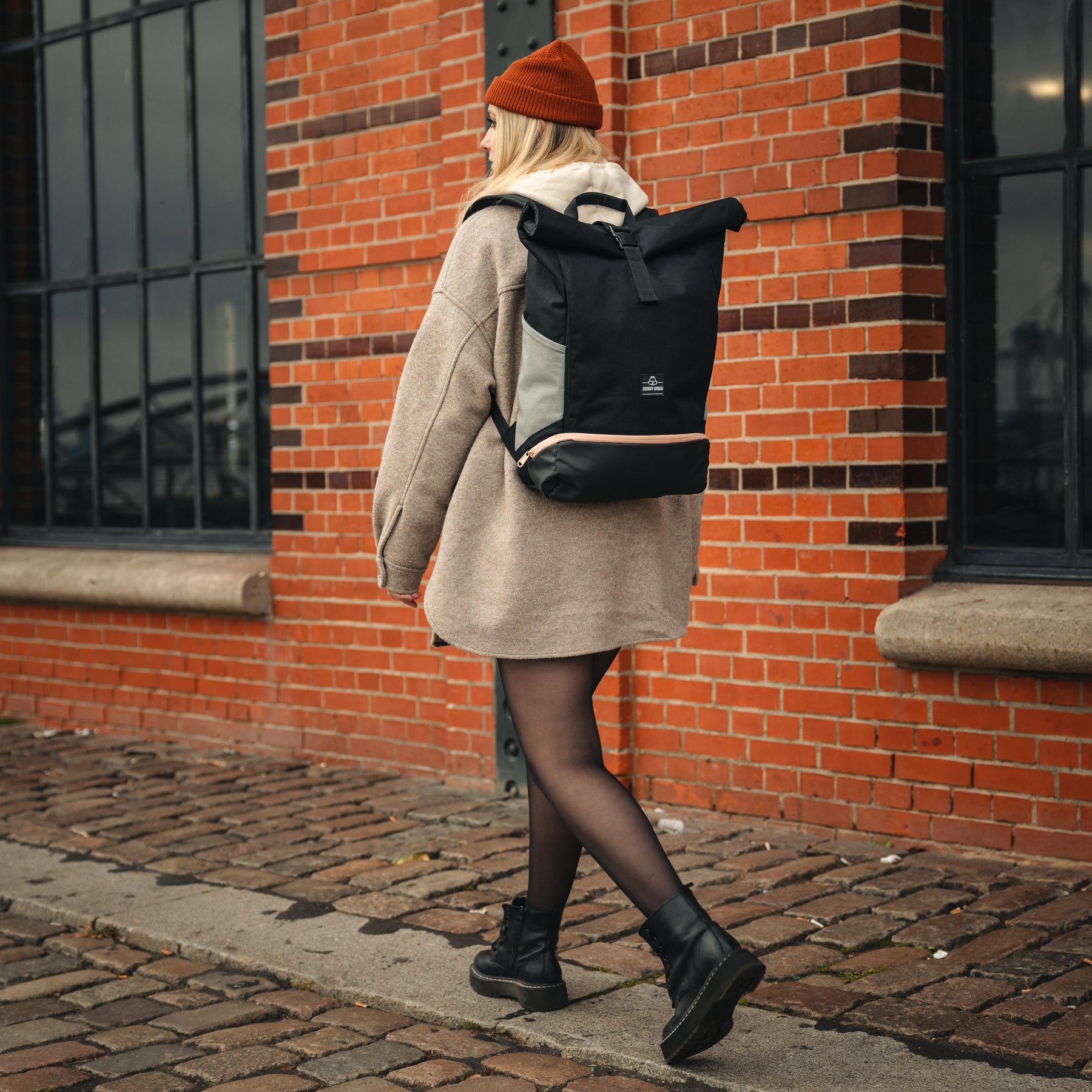 Frau mit Rolltop Rucksack 'Allen Large' von Johnny Urban in Schwarz und Grau, trägt beige Jacke und orange Mütze, geht an roter Ziegelwand