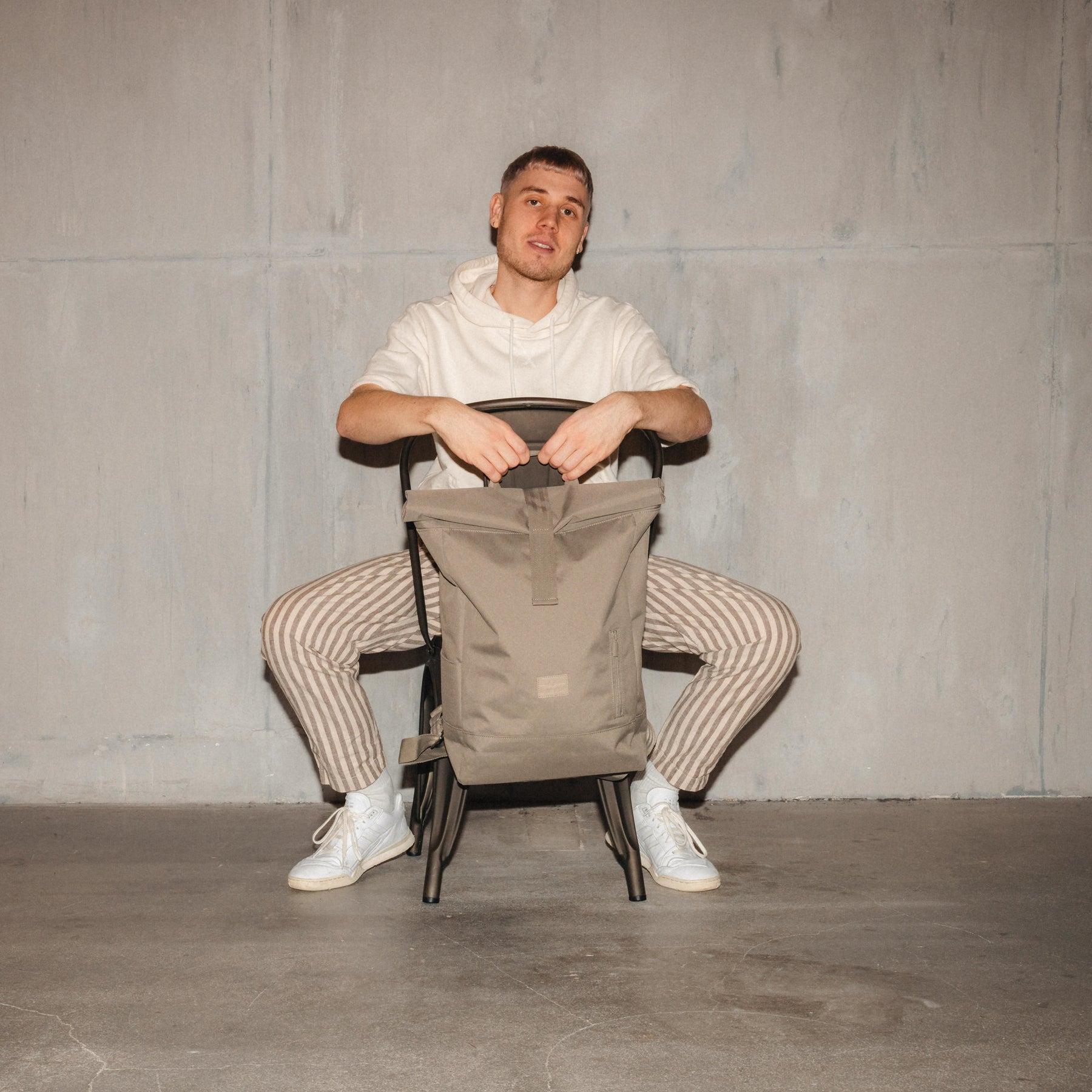 Mann sitzt rückwärts auf Stuhl und hält beigen Rolltop Rucksack 'Robin Large'