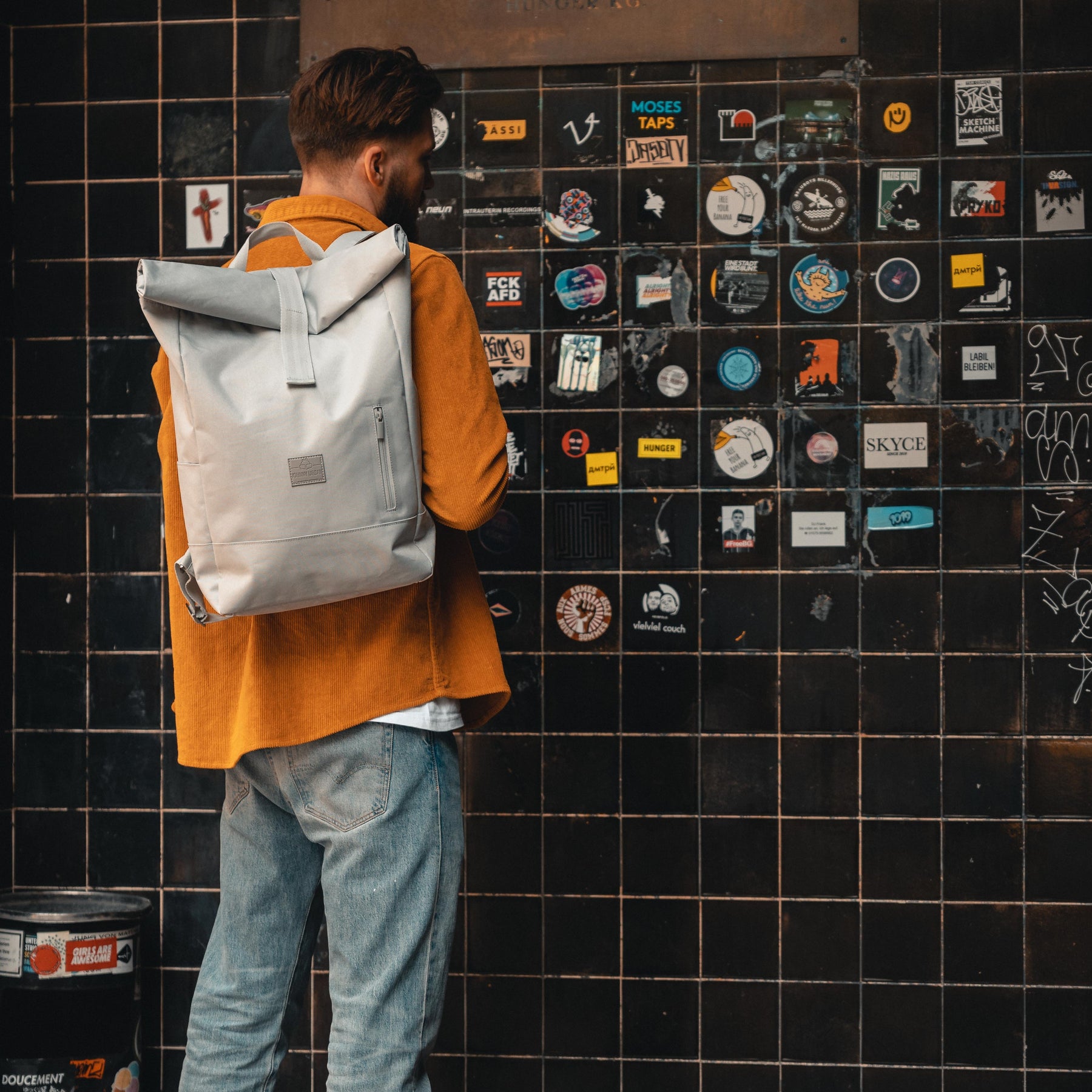 Mann trägt grauen Rolltop Rucksack 'Robin Large' vor schwarzer Wand mit Aufklebern